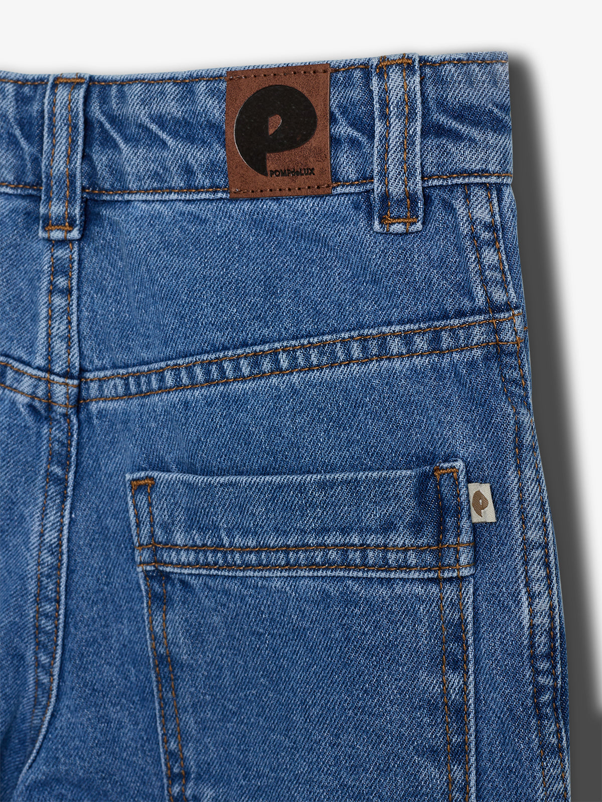 Denim jeans display a rear pocket and belt loops; a brown patch with a black "P" logo above "POMPdelUX" adorns the waistband against a white background.