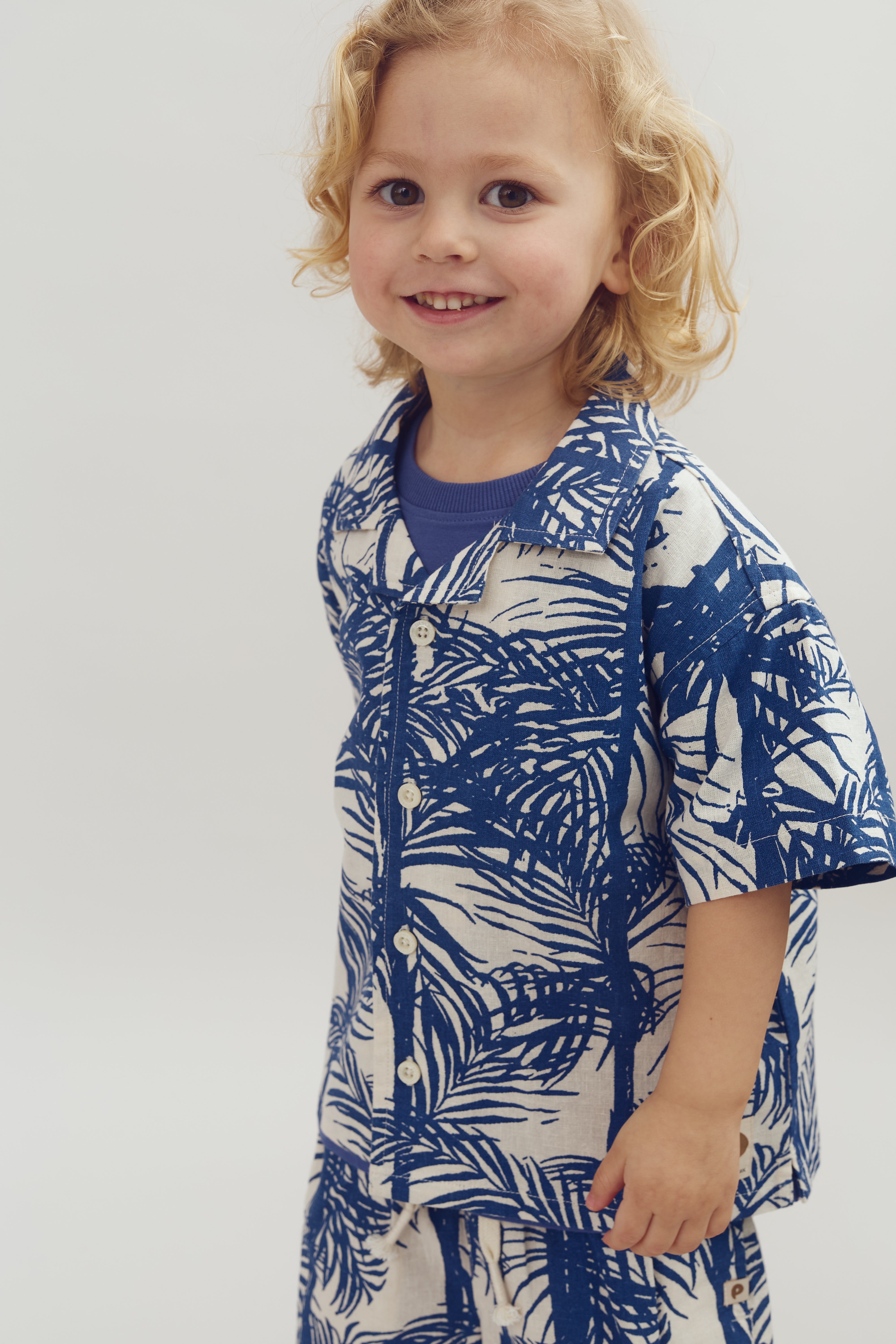A child with curly blonde hair smiles, wearing a blue and white tropical-patterned outfit, in a neutral, softly lit environment.