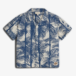 A short-sleeve, button-up shirt features a blue palm tree pattern on a beige background. It includes a small brown patch logo at the lower right corner.