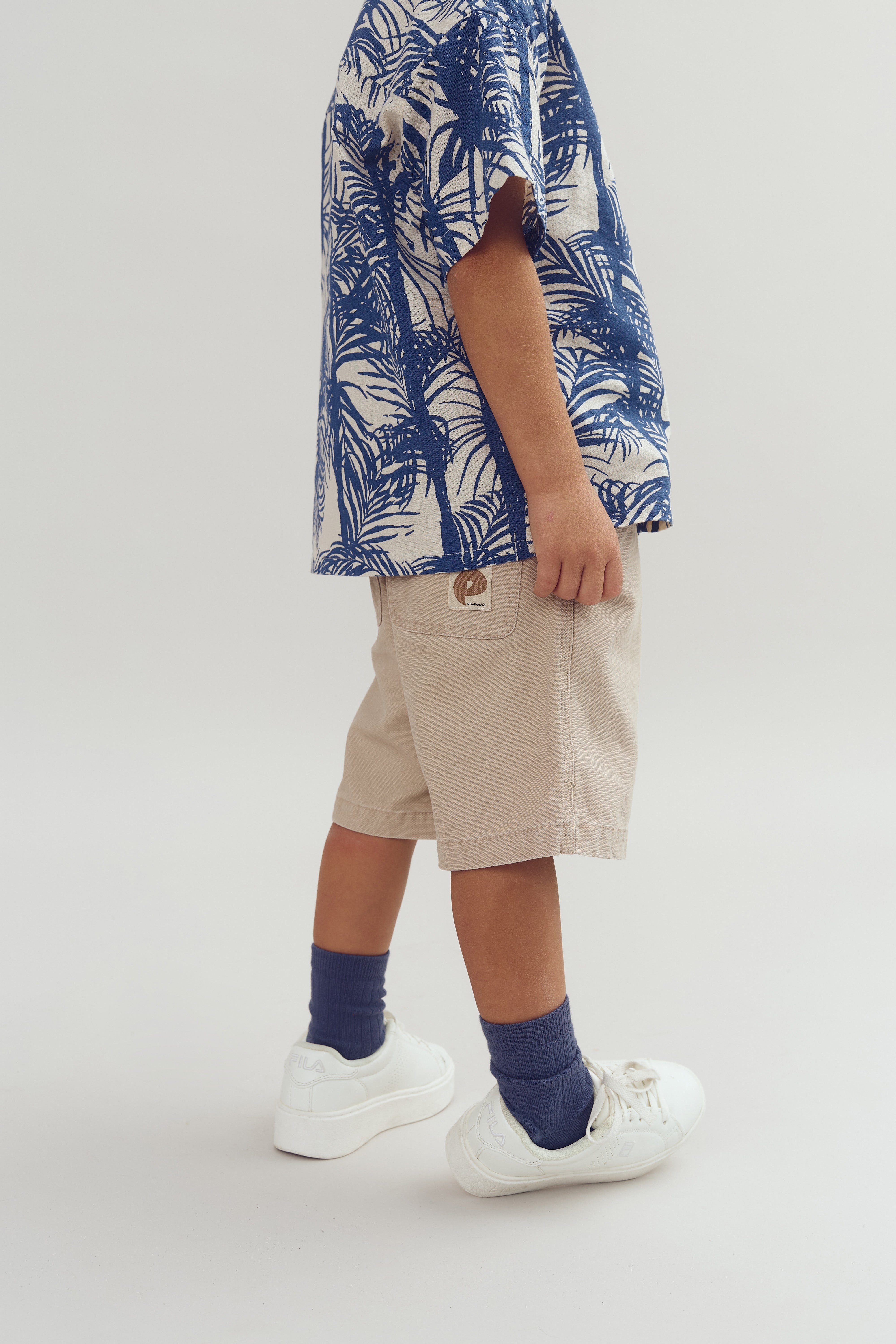 Child wearing blue palm-patterned shirt and beige shorts with a "P" logo, standing on a light surface. Blue socks and white sneakers complete the outfit.