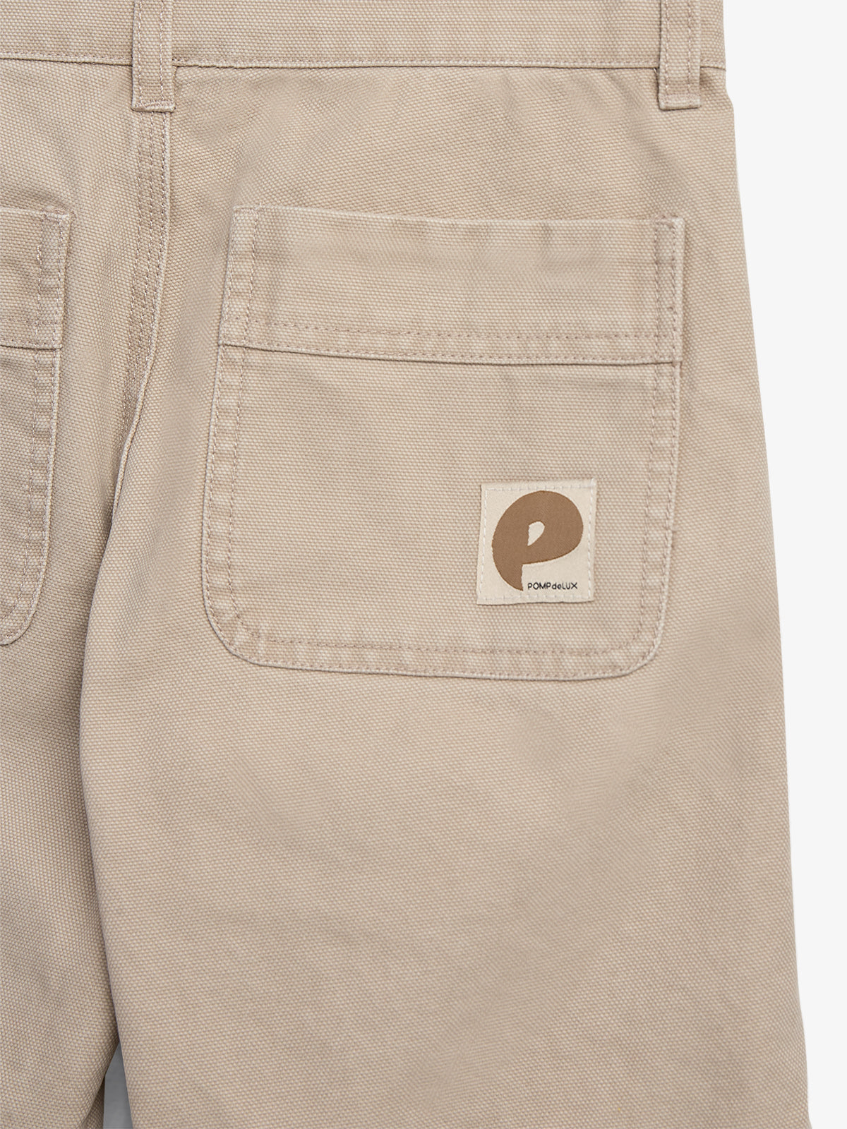 Beige pants feature a back pocket with a beige label showing a brown letter "P" and the text "POMPEUX" underneath, set against a plain background.