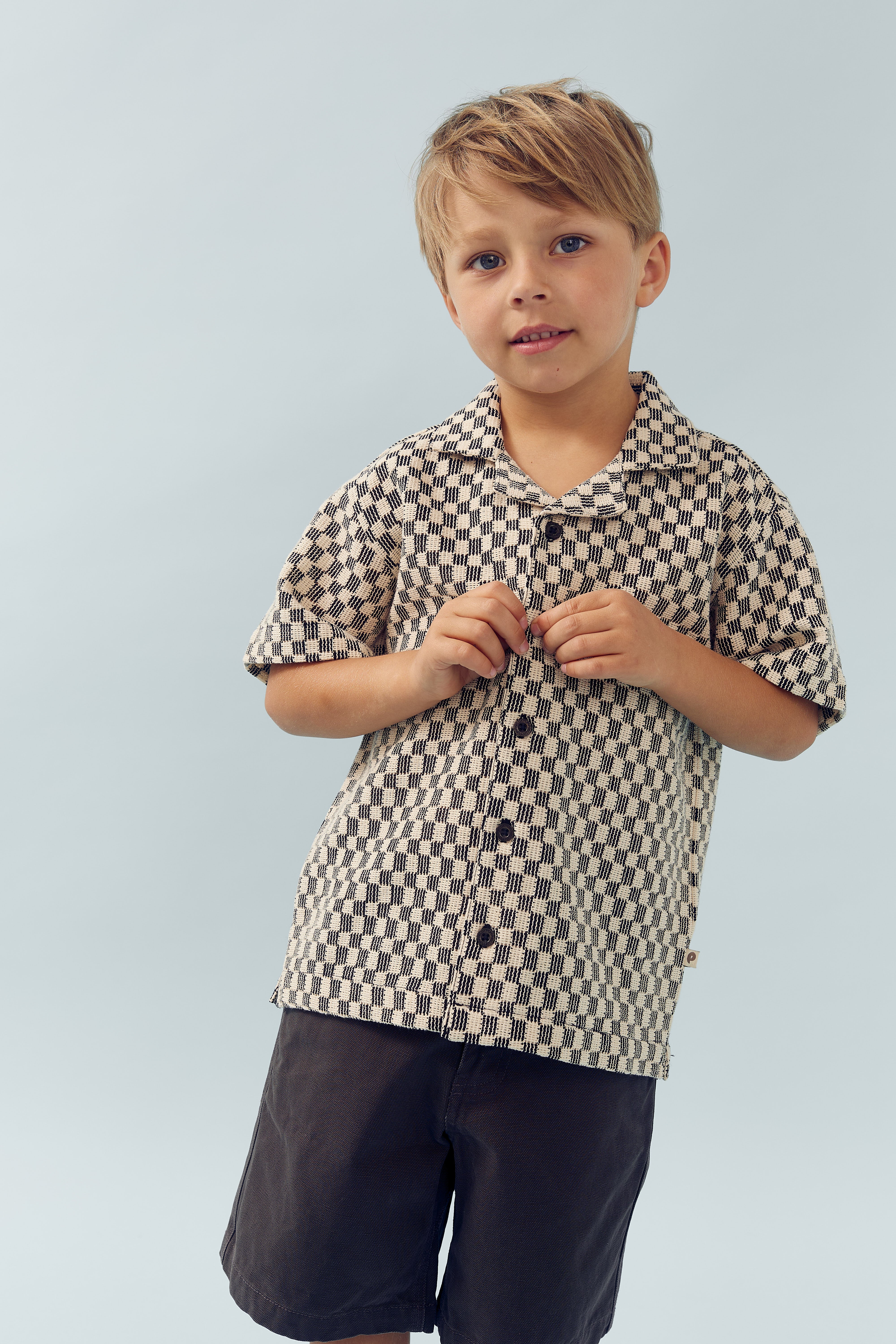 A child wearing a patterned shirt is buttoning it, standing against a plain light blue background. The expression is calm, with hands focused on the task.