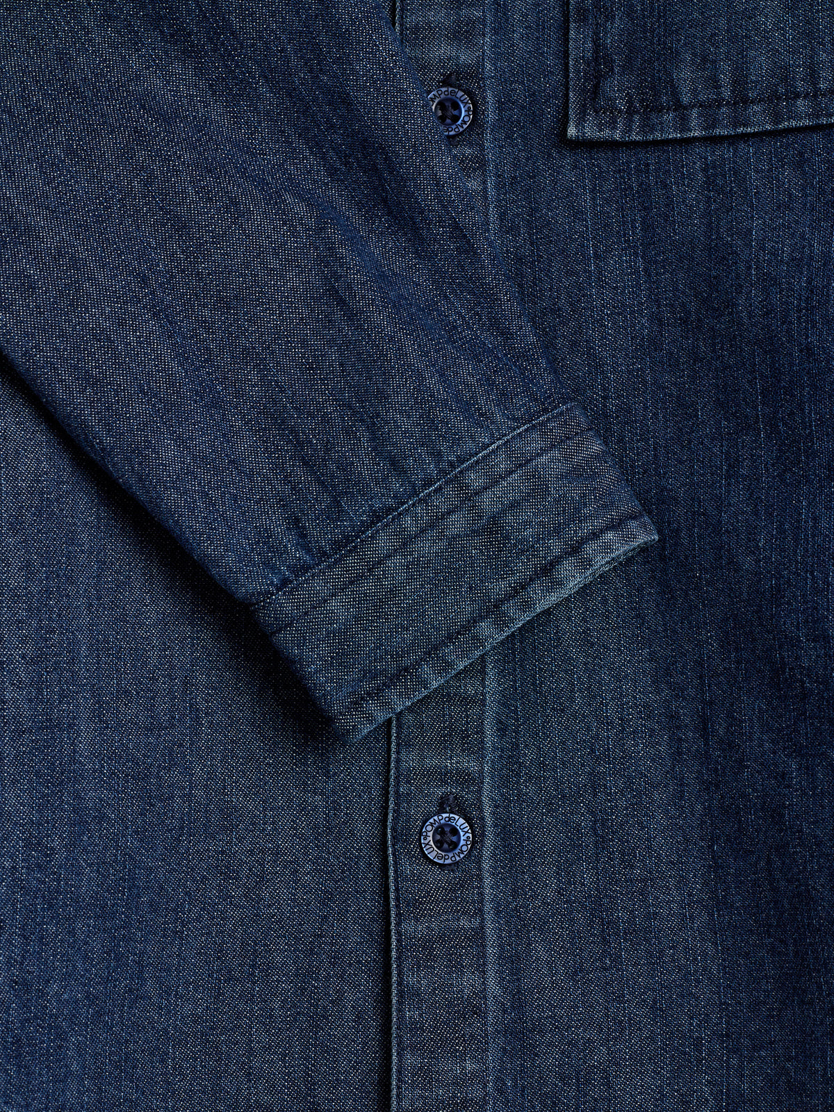 Denim shirt with sleeves folded, positioned over a vertical row of buttons. Two visible buttons are inscribed with "Mode Belgique." The fabric has a dark blue, textured appearance.