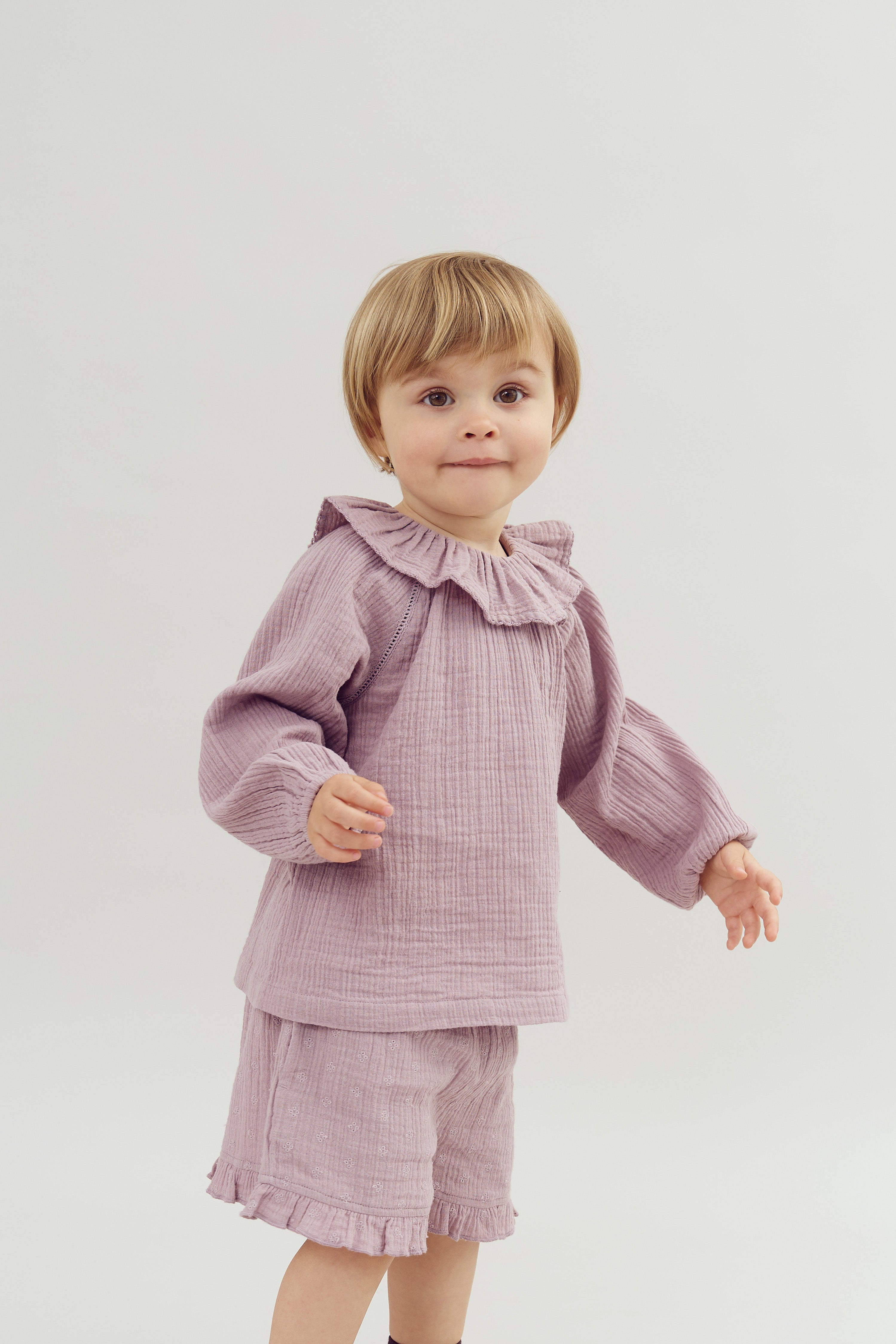 A young child stands wearing a lilac outfit with a ruffled collar, glancing forward, against a plain white background.