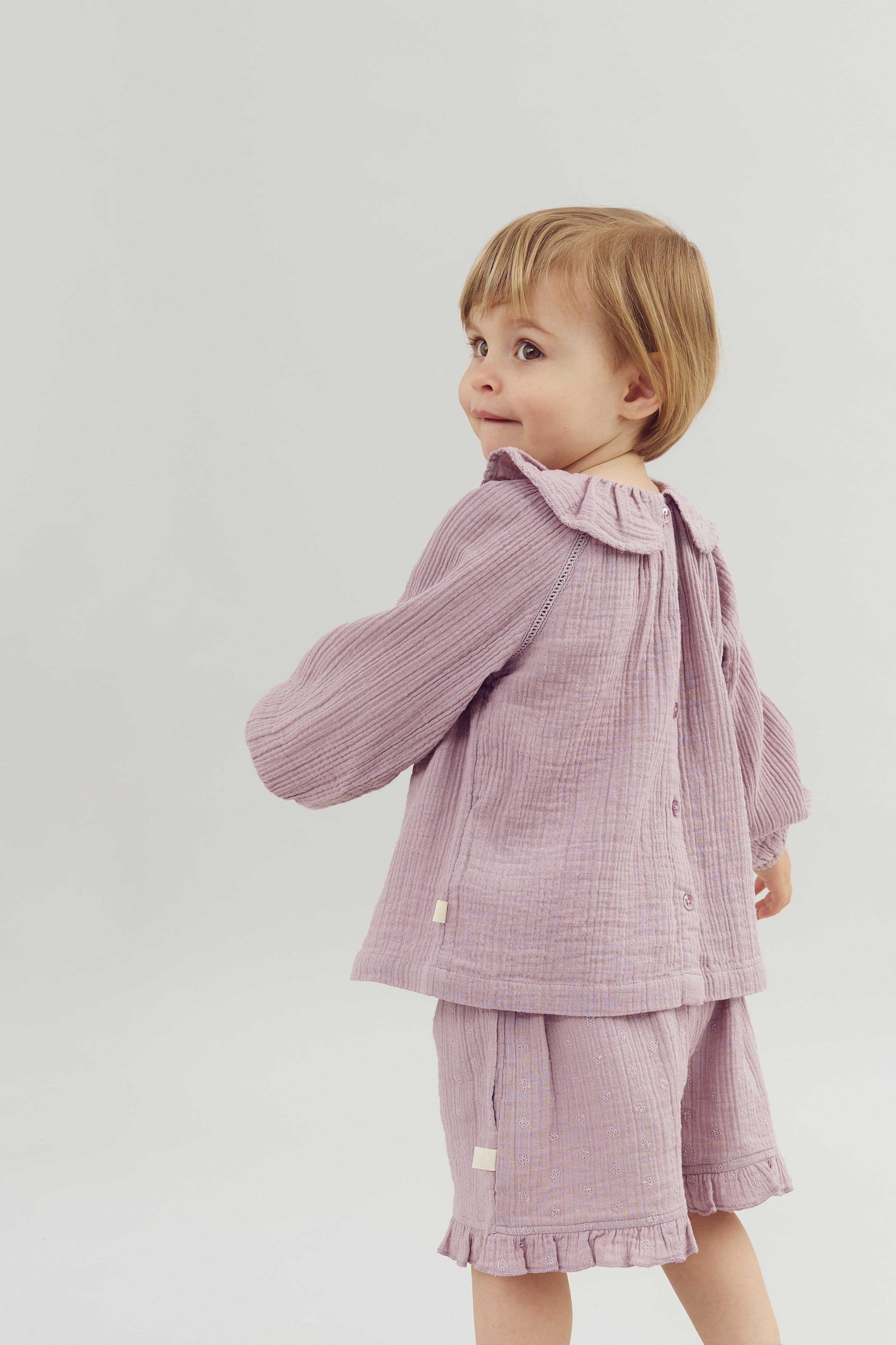 A young child stands turning slightly, wearing a lavender, textured two-piece outfit with a ruffled collar and hem, against a plain white background.