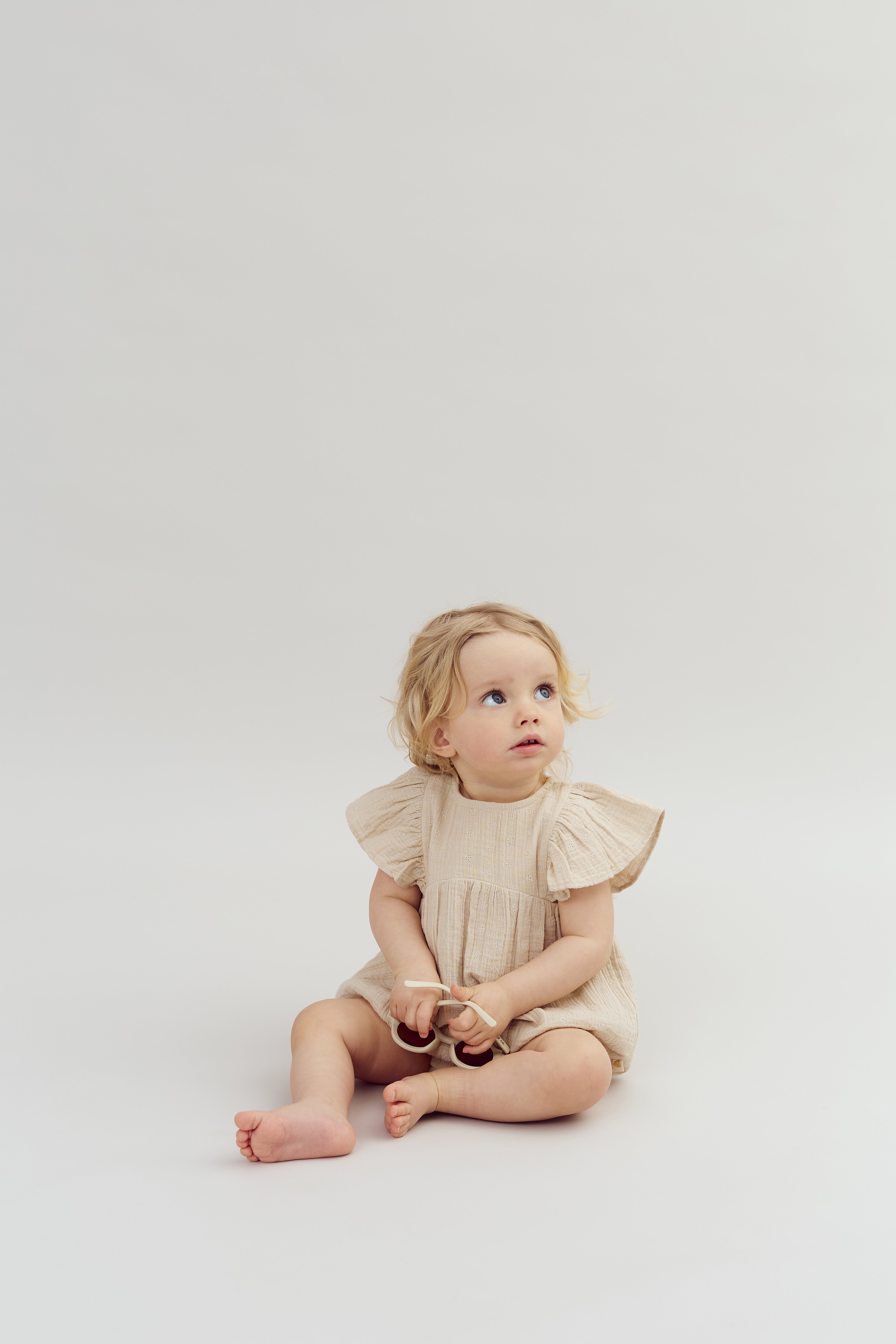 A toddler sits on a light background, wearing a beige dress, holding a toy, and looking upward with an inquisitive expression.