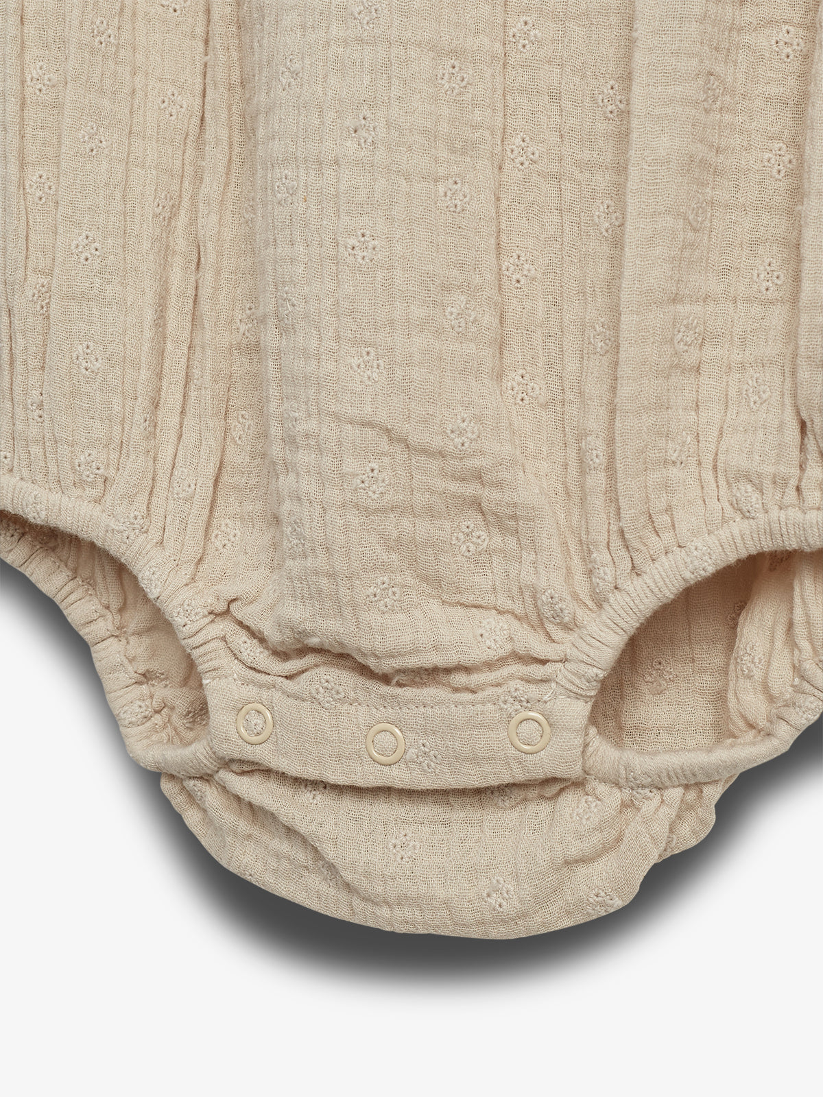 Beige baby romper with small embroidered patterns and snap buttons for closure, lying against a plain white background. The fabric is textured with tiny floral designs.