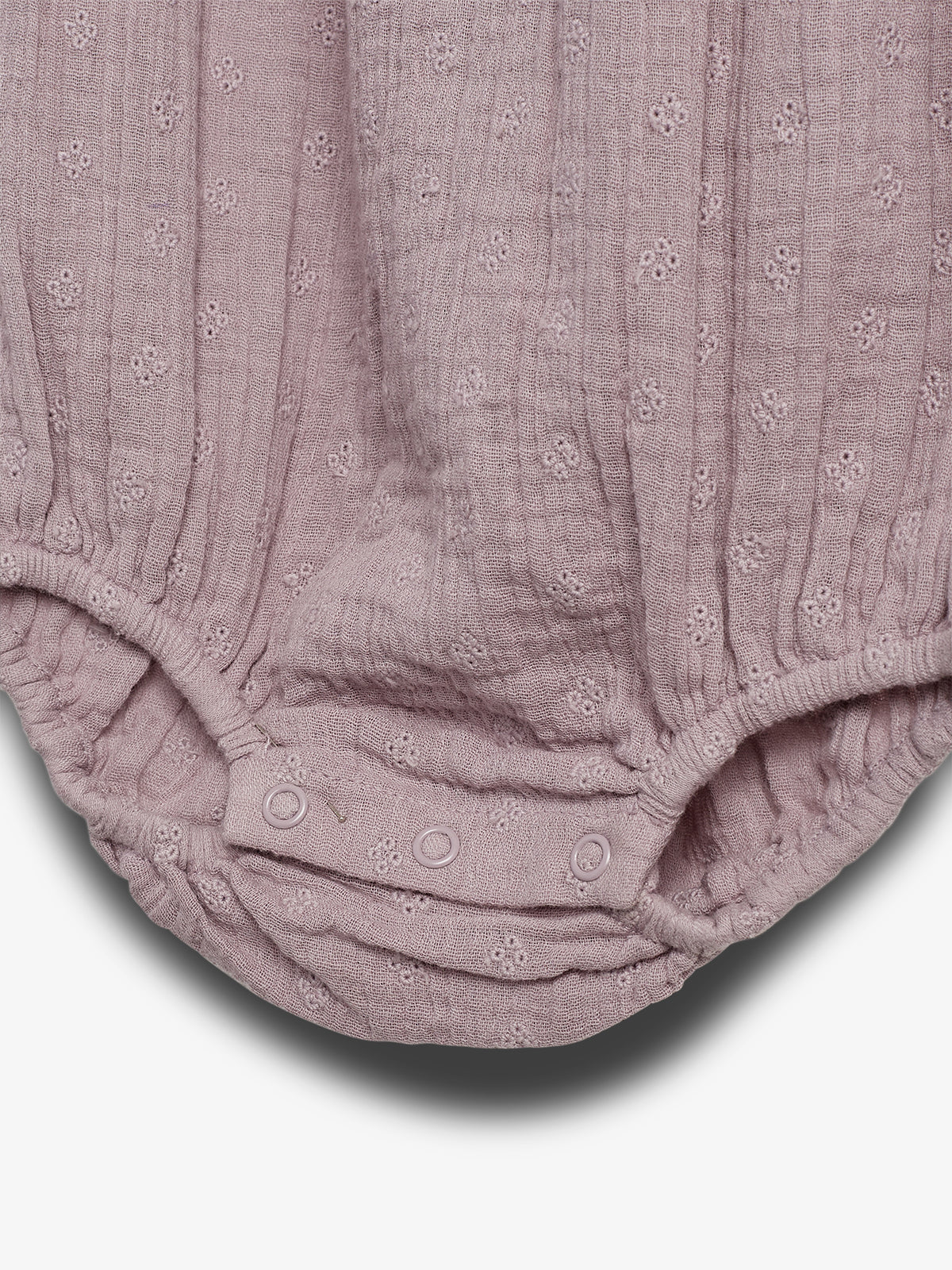A light purple fabric with small floral patterns, featuring buttons and elastic edges. It seems to be the bottom portion of a baby romper set against a white background.
