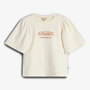 Cream-colored T-shirt featuring puffed sleeves, displays text "AMOUR" and "J’ADORE LE SOLEIL." The shirt is laid flat against a plain white background.