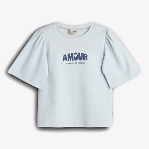 A short-sleeved, light blue shirt with puffed sleeves displays the text "AMOUR" in bold, large letters and "J'ADORE LE SOLEIL" beneath. The shirt is on a plain background.