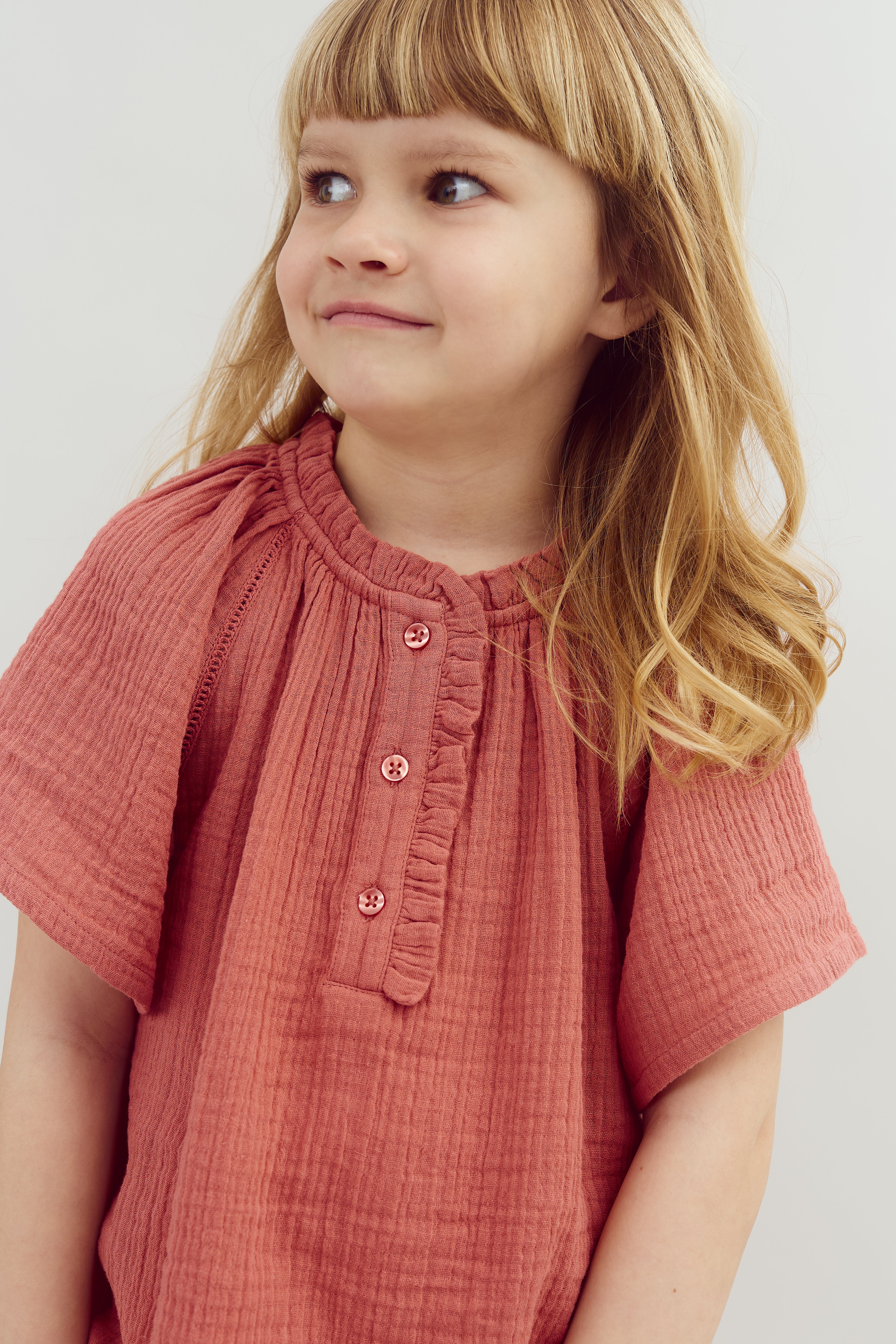 A child with long hair wears a pink button-up top, looking to the side with a slight smile, set against a plain background.