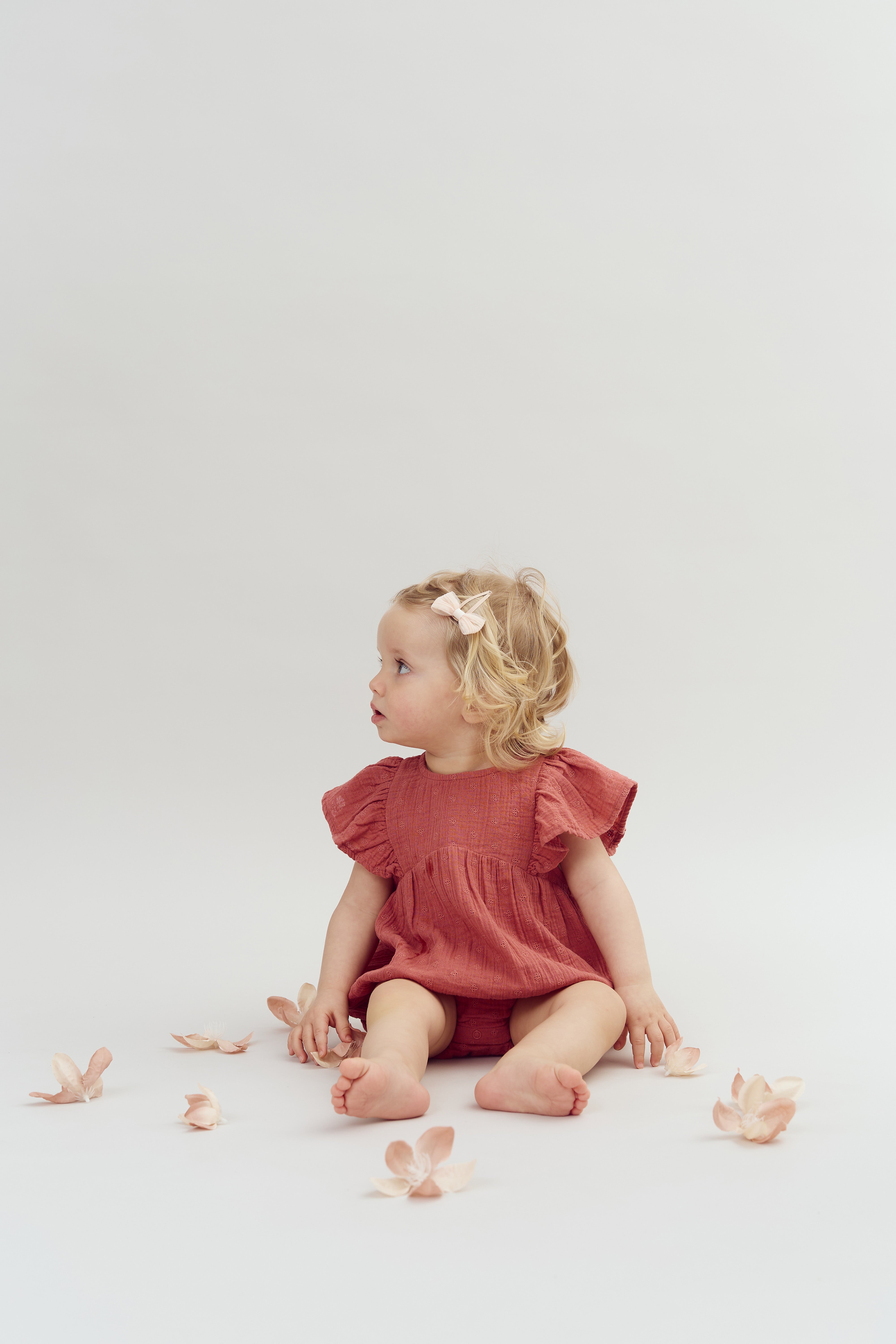 A young child sits on the floor, wearing a red dress and a hair bow, surrounded by scattered, pale flowers against a plain background.