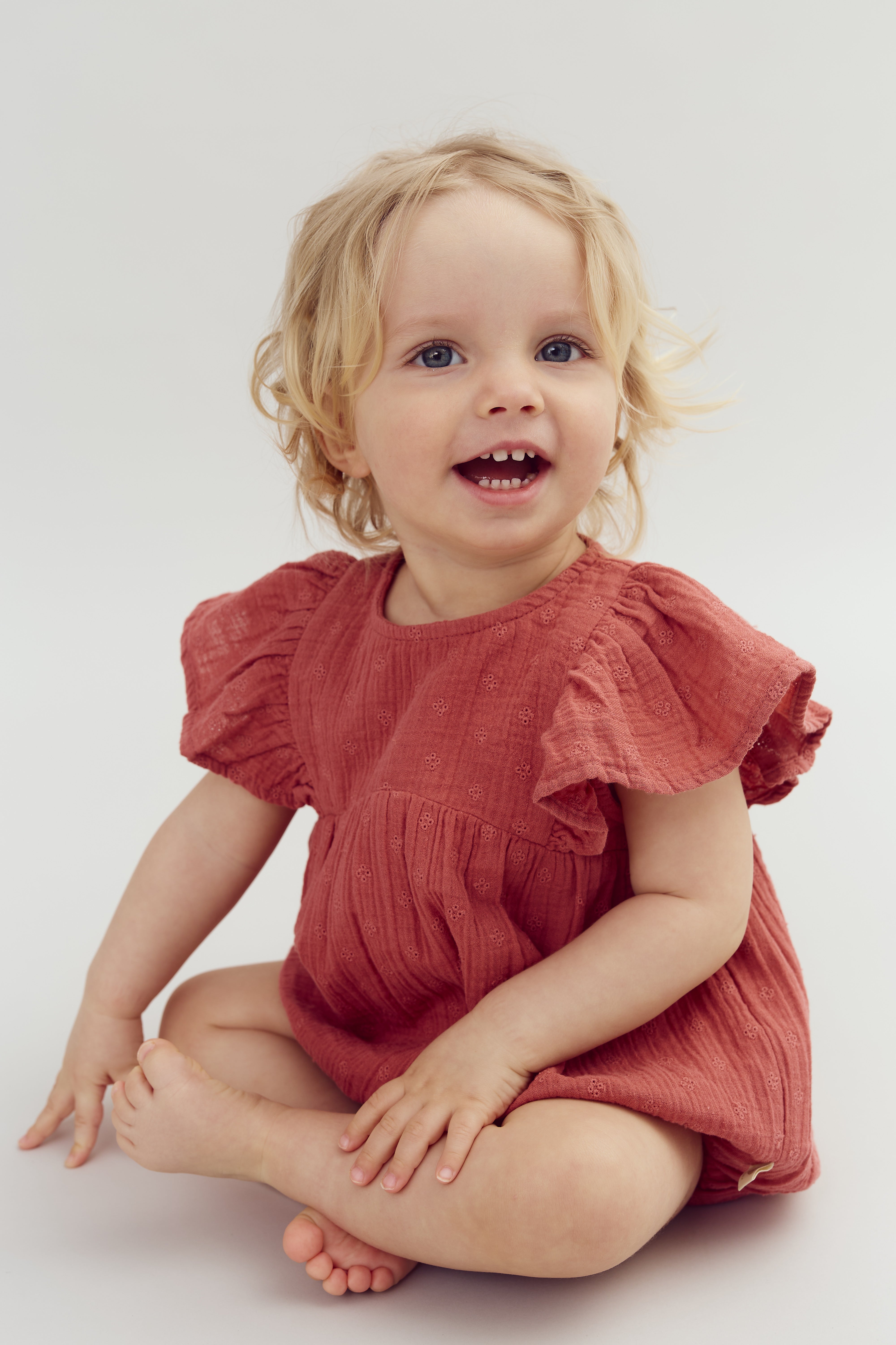 A toddler in a rust-colored dress sits cross-legged, smiling brightly. The background is a neutral, light gray, creating a clean and simple setting.