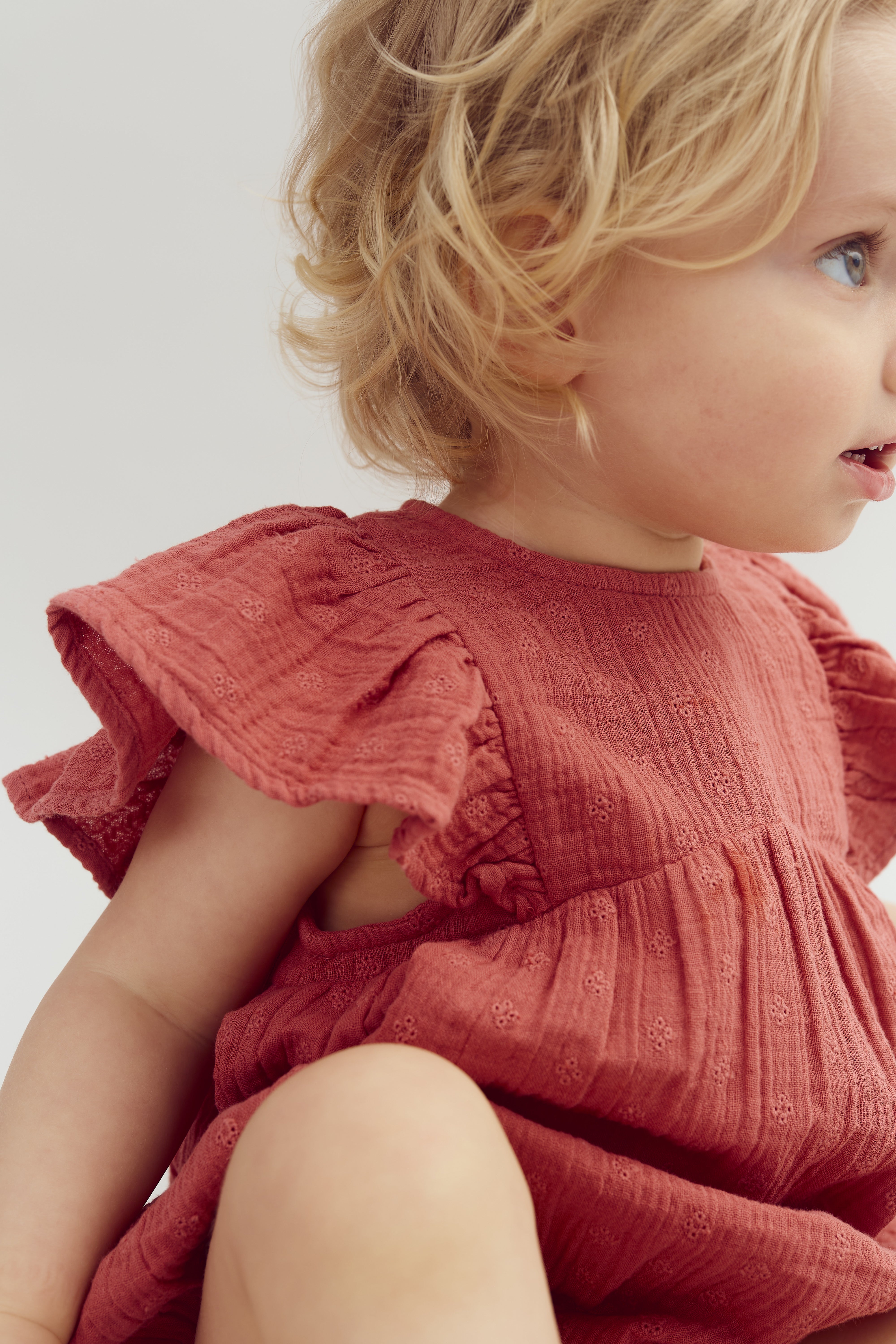 A toddler wearing a textured, red dress looks to the side with soft, wavy blonde hair. The background is plain and light, emphasizing the child’s profile.