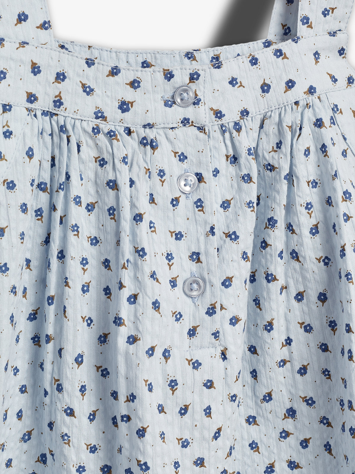 Light blue fabric with a pattern of small blue flowers and brown leaves, featuring three white buttons, is gathered and pleated, suggesting a garment, in a neutral lighting setting.