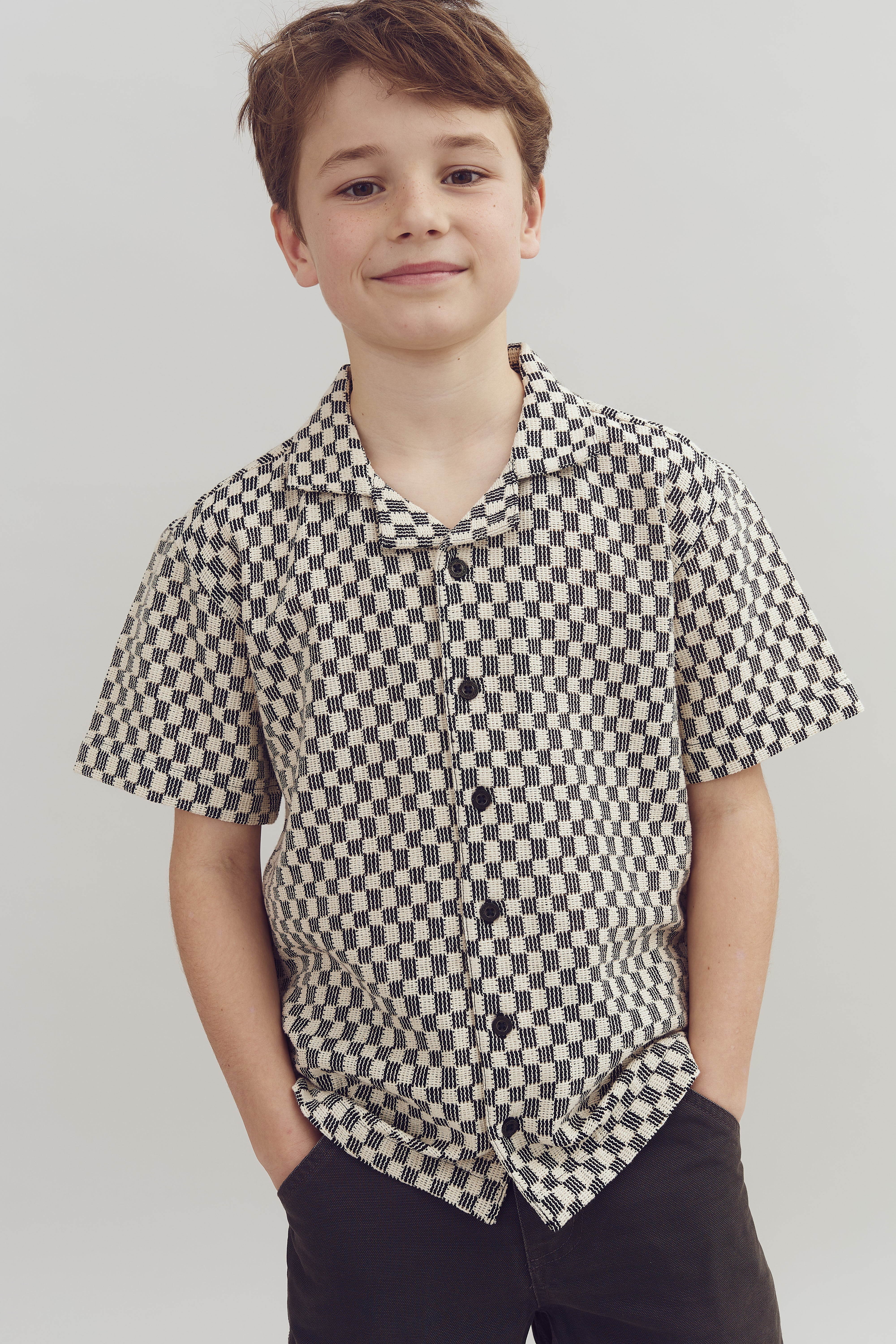 Boy wearing a checkered shirt smiles with hands in pants pockets, standing against a plain light background.