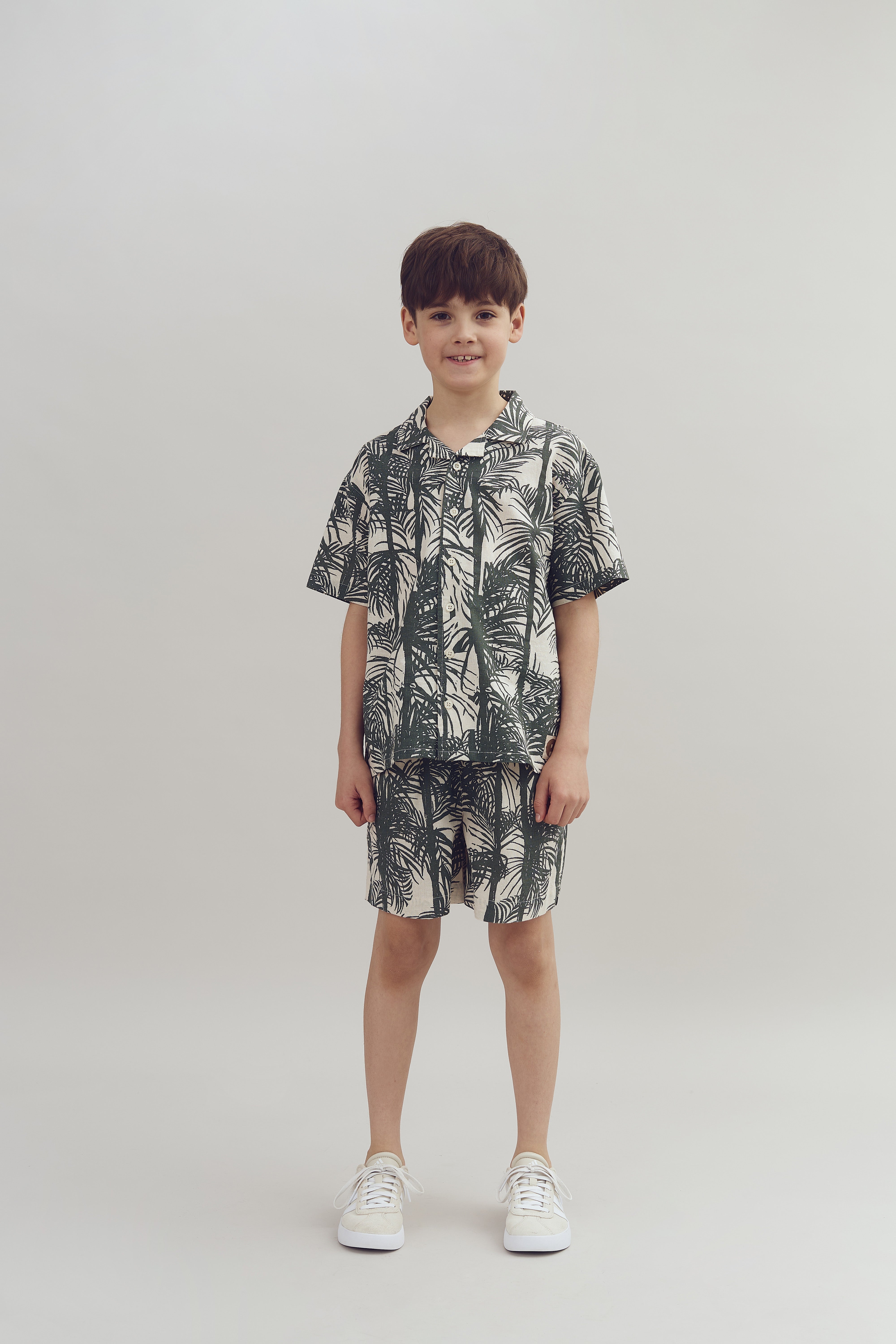 A child stands smiling, wearing matching leaf-patterned shirt and shorts with white sneakers, set against a plain, light gray background.