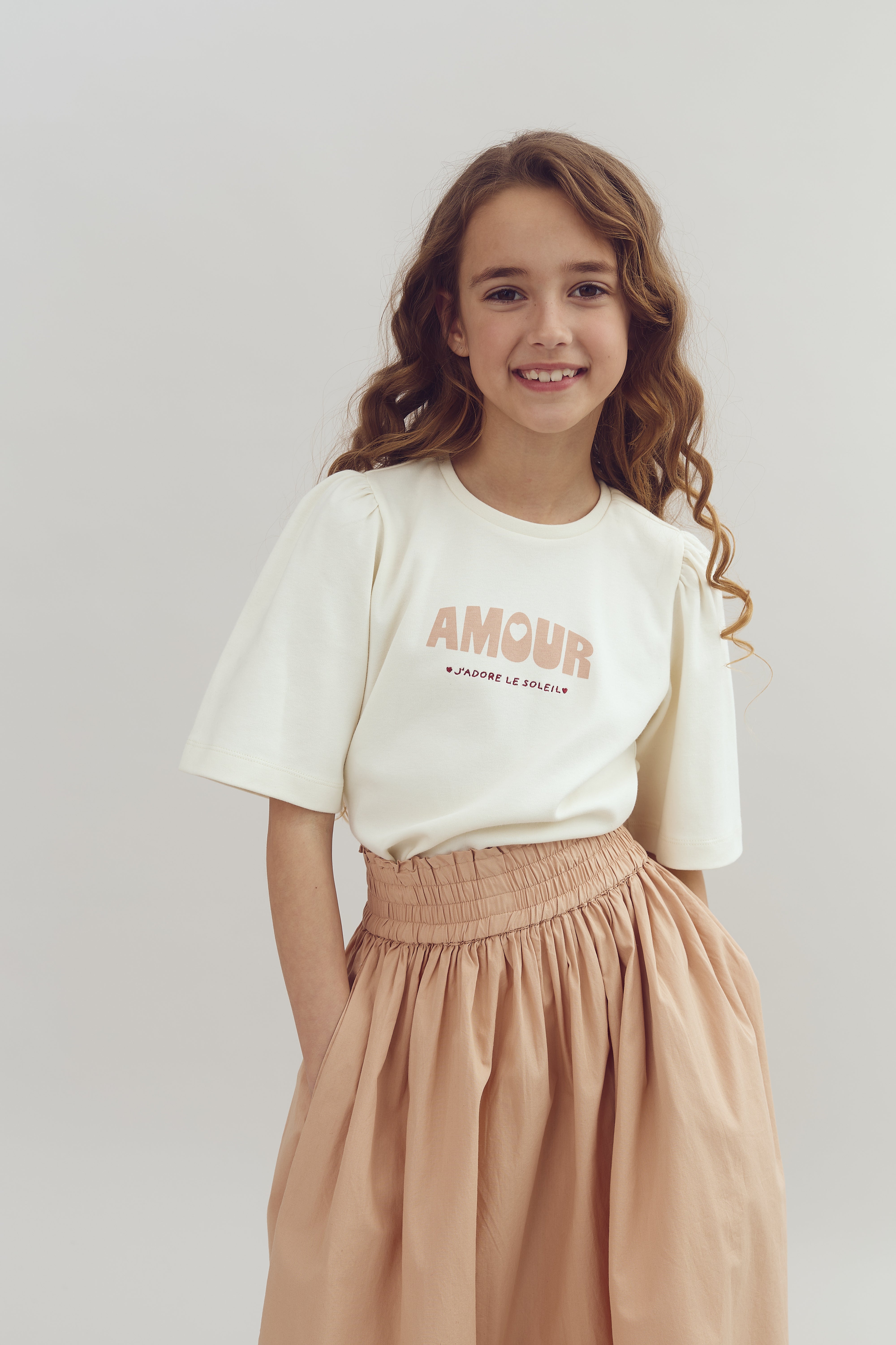 A smiling girl stands with hands in pockets, wearing a white shirt with "AMOUR J'ADORE LE SOLEIL" printed on it, and a beige skirt. She is in a neutral, light background.