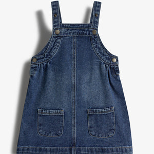 Denim pinafore dress hangs against a plain white background, featuring adjustable straps with buttons and two front pockets. The dress has a simple, casual design.