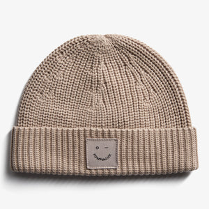 A beige knit beanie with a folded brim features a small patch showing a simple face and the text "POMPdeLUX." It's displayed against a plain white background.