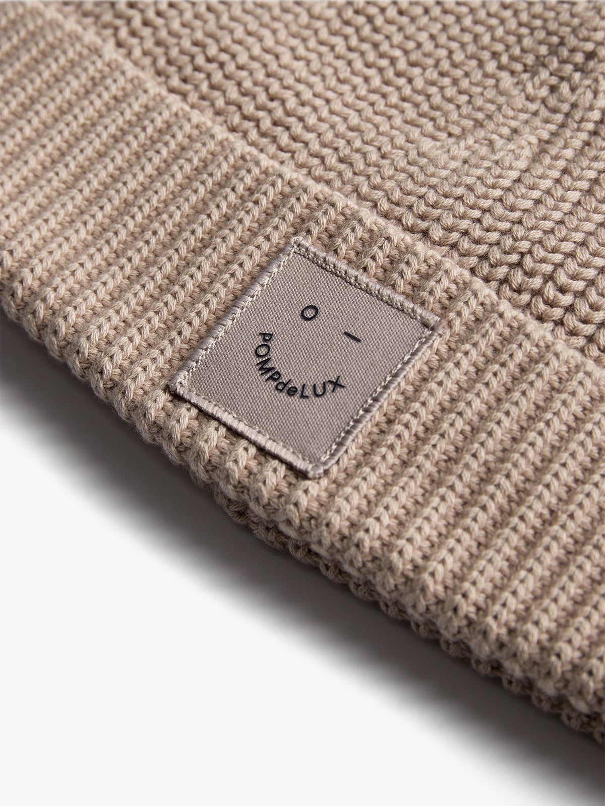 A beige knitted beanie features a small fabric patch displaying a simple face design and the text "POMPdeLUX" against a white background.