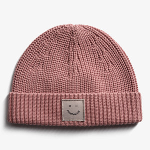 A pink, ribbed knit beanie with a folded brim features a small square patch displaying a simple face and the text "POMPdeLUX" against a plain white background.