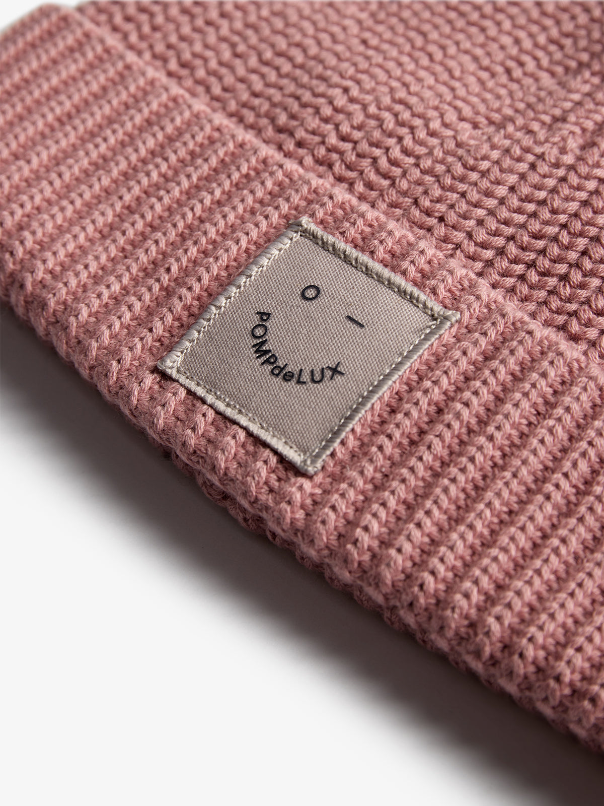 A pink knitted beanie features a labeled patch. The patch displays a minimal face and the text "POMPdeLUX." The background is a plain, light surface.