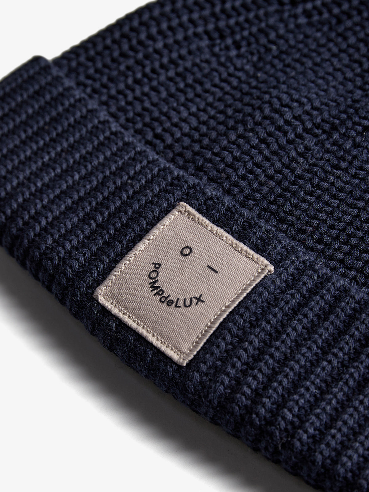 A navy blue knit hat features a light-colored patch with a simple face and the text "POMPdeLUX." The hat lies flat against a white background, emphasizing its texture.