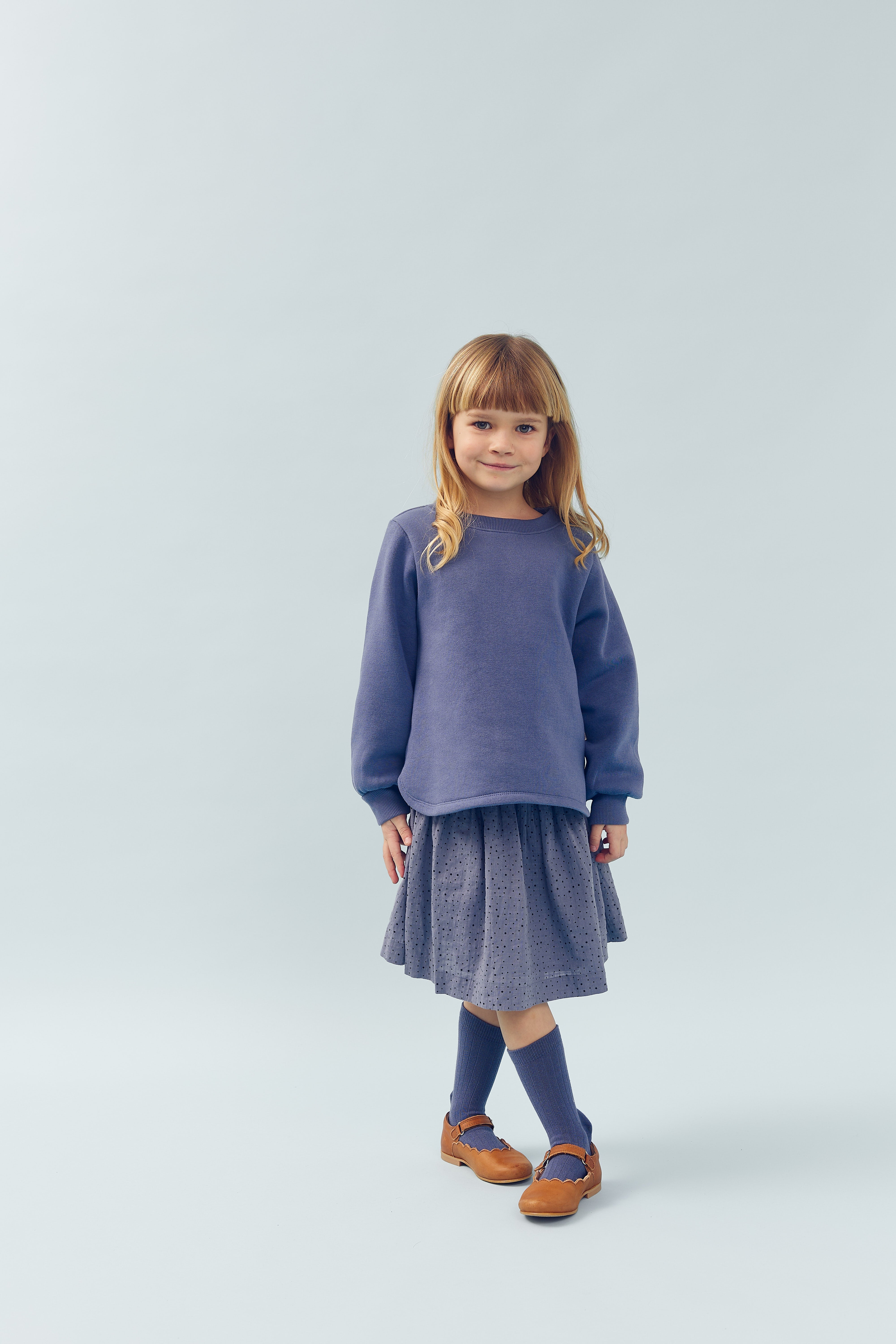 A young girl stands smiling, wearing a blue sweater, skirt, matching socks, and brown shoes, against a light blue background.