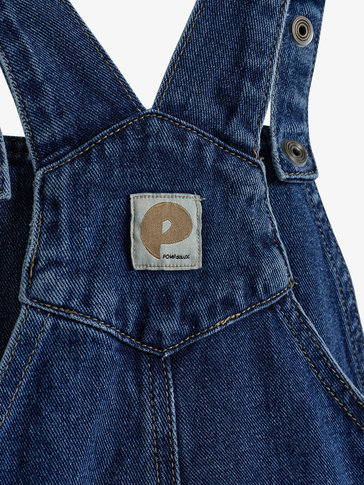 Denim overalls display a rectangular patch with a brown circle and text "POMPEALUX," featuring metal fasteners. The fabric appears dark blue with stitching detail, suggesting casual clothing.