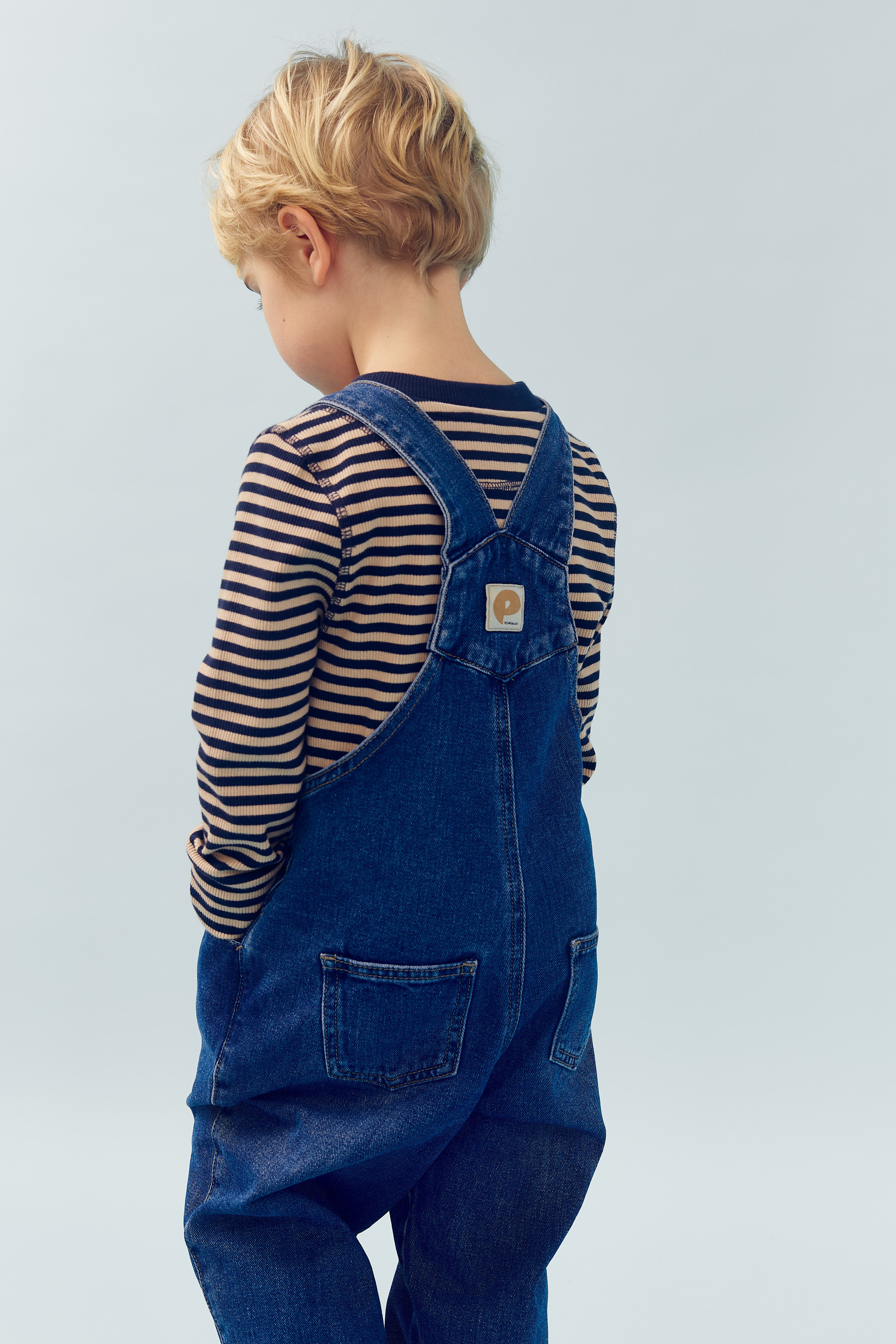 A child stands with their back turned, wearing blue denim overalls and a striped shirt, in front of a plain light blue background.