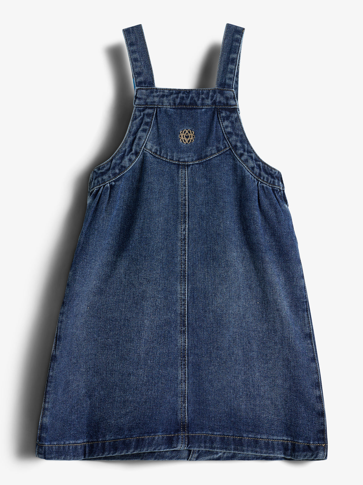 A denim pinafore dress hangs against a plain white background, featuring thick straps and a small, embroidered circular pattern on the chest.