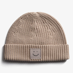 A beige knitted beanie displaying a simple, embroidered smiley face patch with text "POMPdeLUX" on its fold-up edge, against a plain white background.