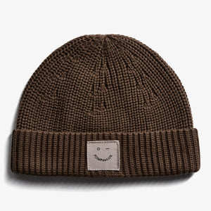 A brown knit beanie lies flat on a white background. It features a folded brim and a patch with a smiley face and the text "POMPdeLUX" on the front.