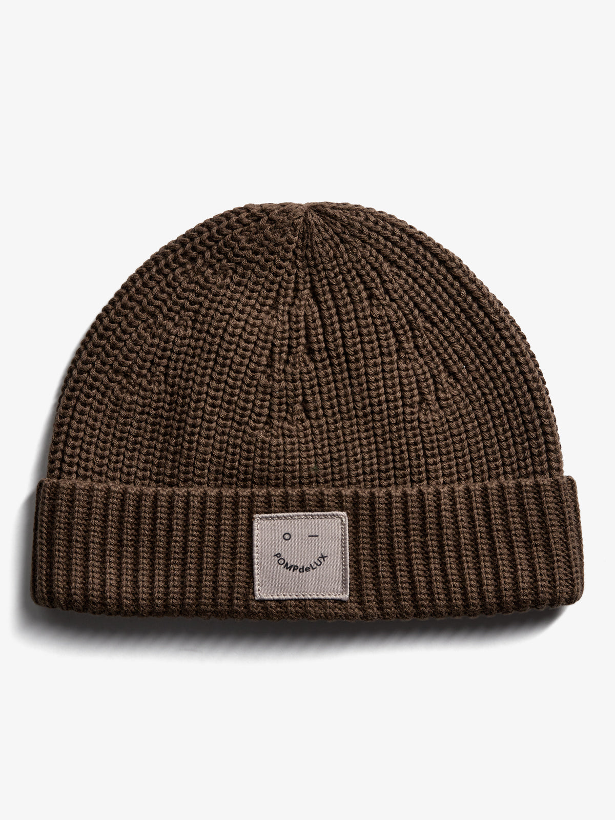 A brown knit beanie lies flat on a white background. It features a folded brim and a patch with a smiley face and the text "POMPdeLUX" on the front.