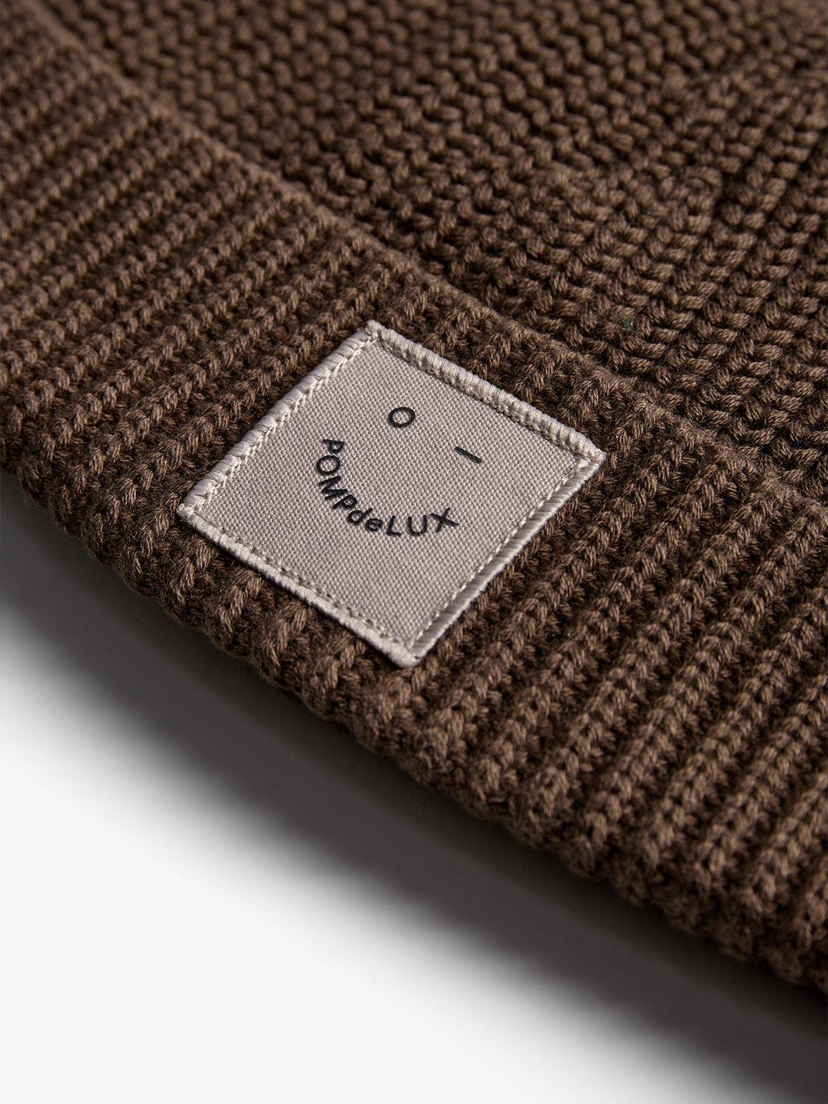 Brown knitted beanie featuring a rectangular label. The label shows a simple face design and the text "POMPdeLUX." The scene highlights the texture and craftsmanship of the fabric.