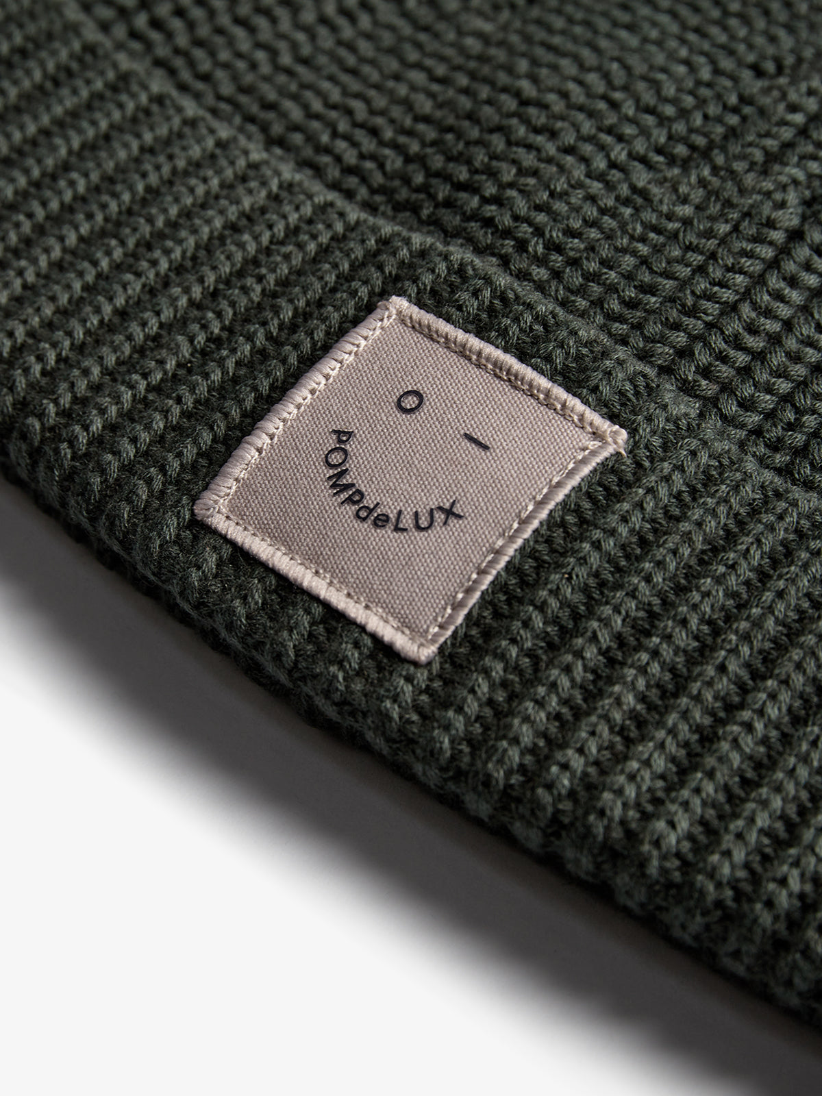 A dark knitted beanie displays a fabric patch with a simple face design and the text "POMPdeLUX" stitched onto it, resting on a light gray background.