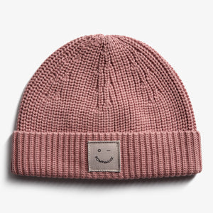 A pink knitted beanie with a folded brim features a stitched patch displaying an abstract face and the text "POMPdeLUX." The beanie rests on a plain white background.