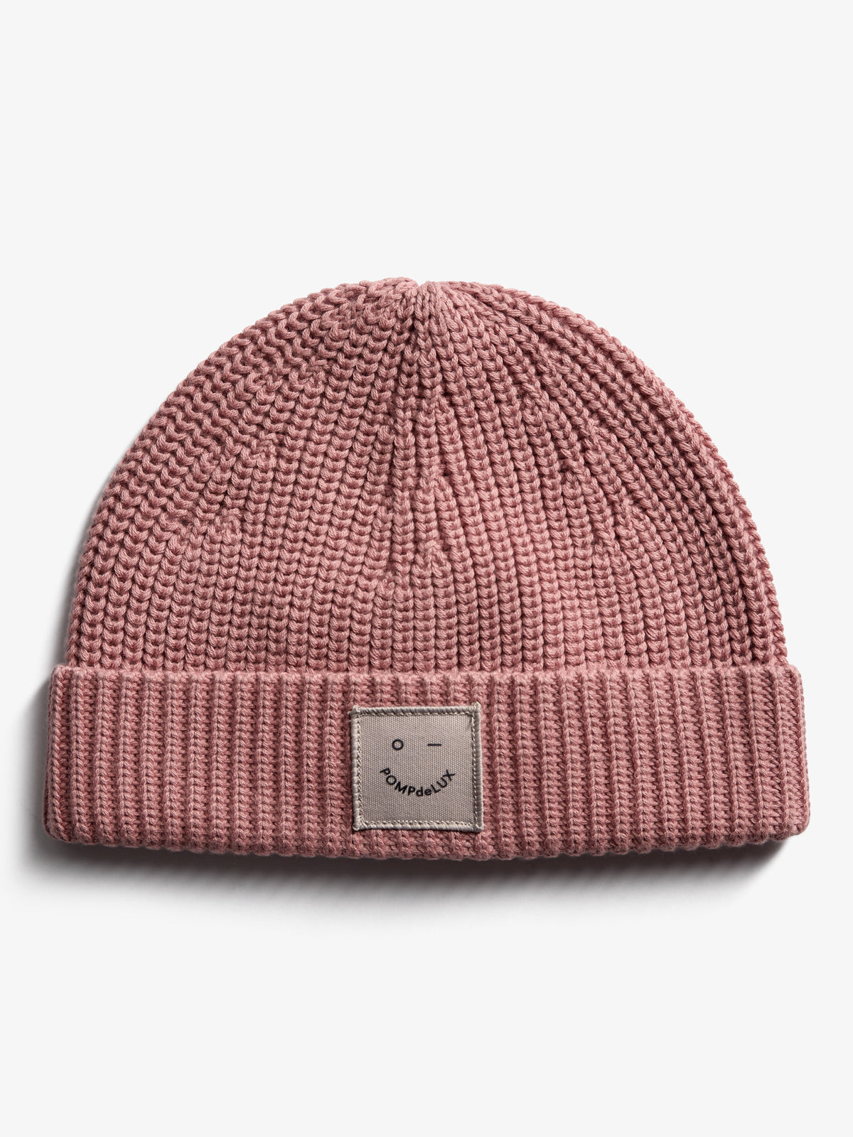 A pink knitted beanie with a folded brim features a stitched patch displaying an abstract face and the text "POMPdeLUX." The beanie rests on a plain white background.