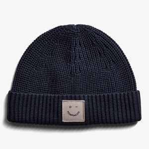 A navy blue knit beanie with a folded brim, featuring a small beige patch displaying a minimalist face and the text "POMPdeLUX" on a plain white background.