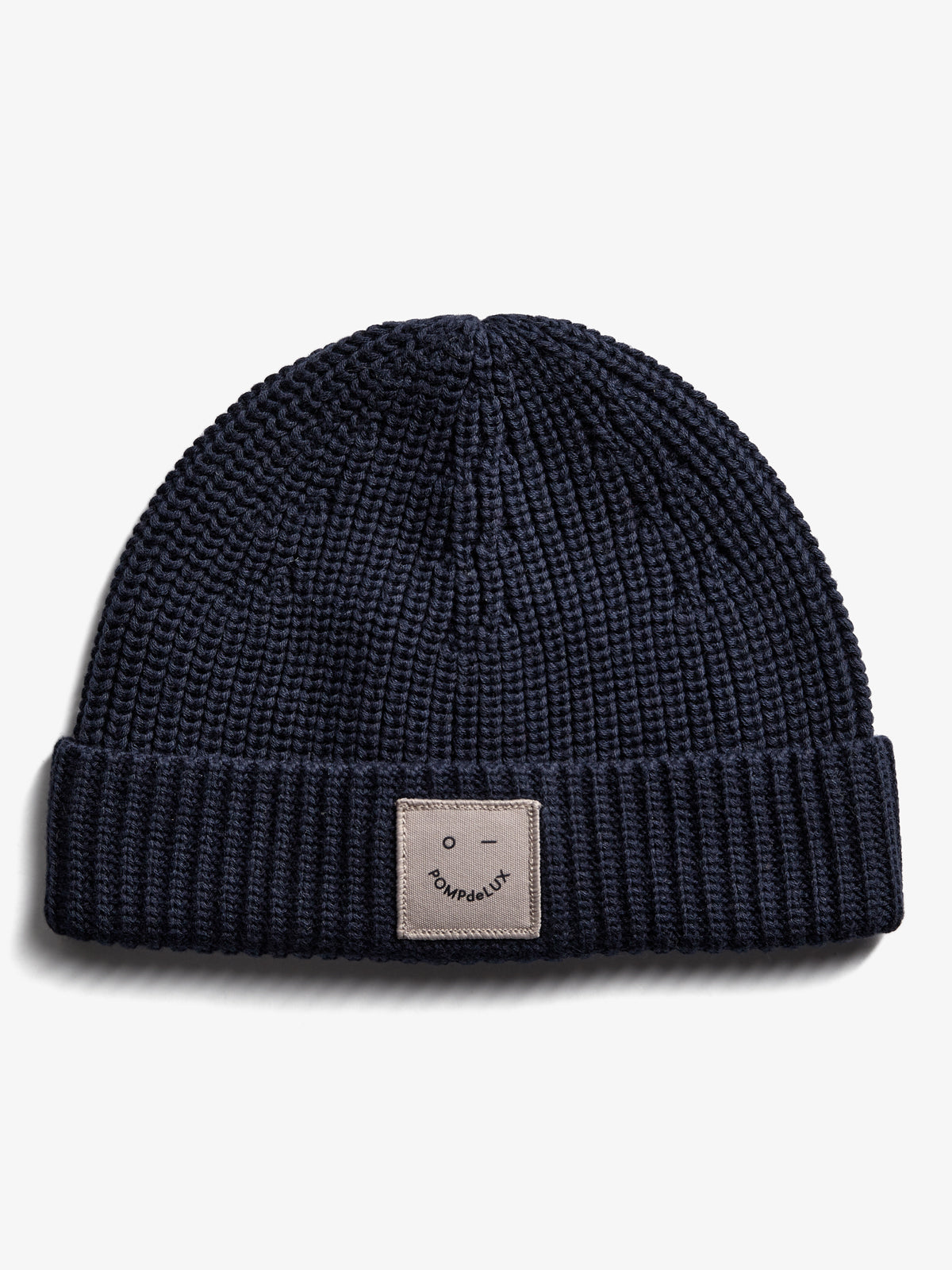A navy blue knit beanie with a folded brim, featuring a small beige patch displaying a minimalist face and the text "POMPdeLUX" on a plain white background.