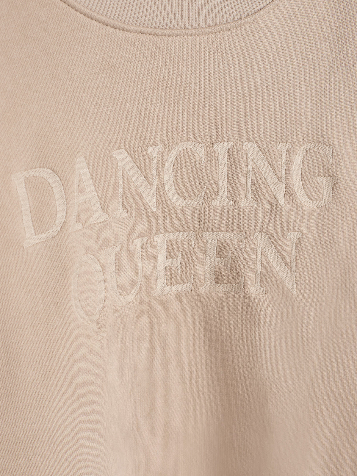 A beige sweatshirt features the embroidered phrase "DANCING QUEEN" in large, bold letters, centered on the chest against a plain background.