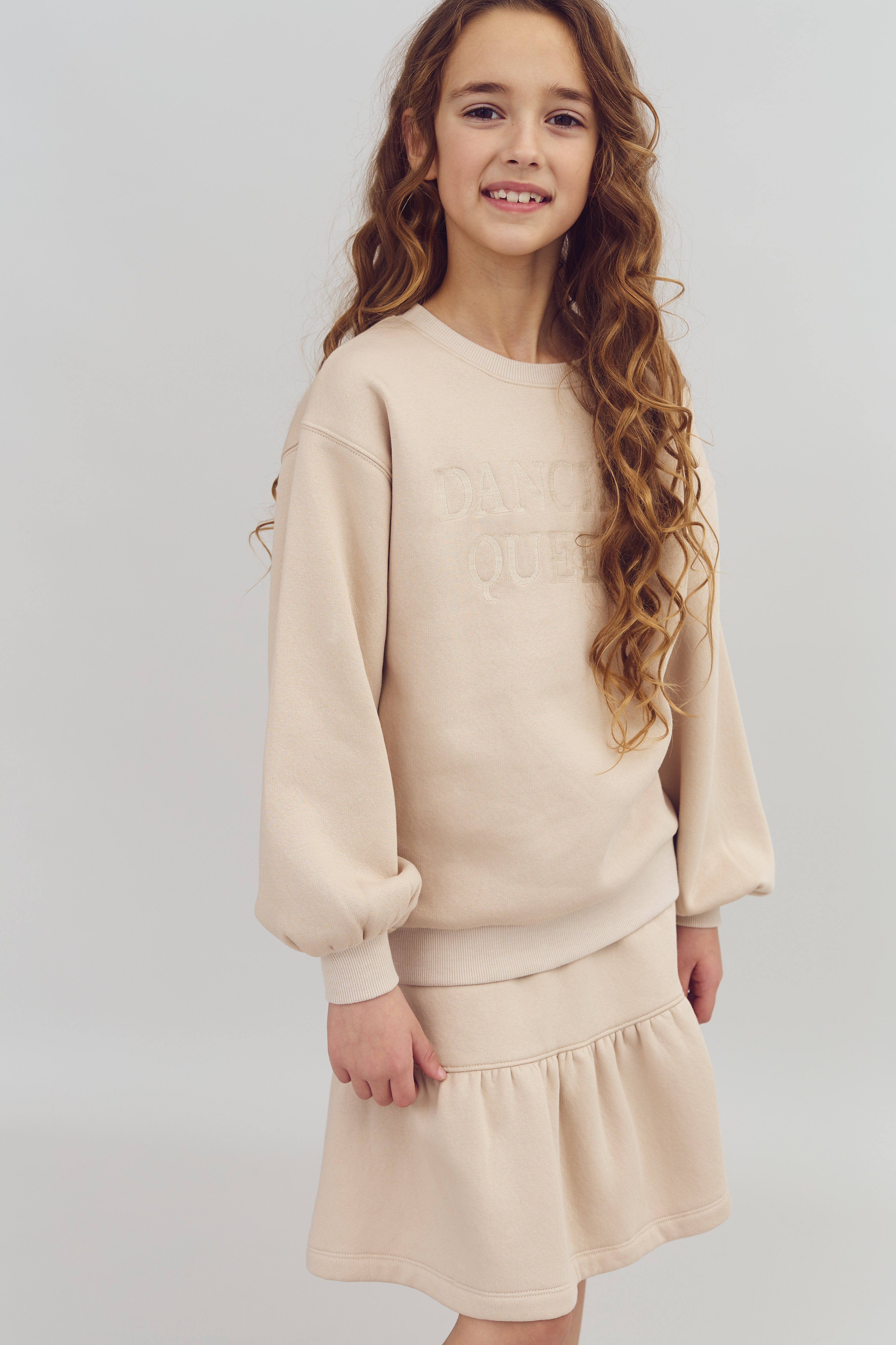 A young girl stands smiling, wearing a beige sweater with "DANCING QUEEN" embossed, and a matching skirt. Her long, wavy hair cascades down. She is against a plain gray background.