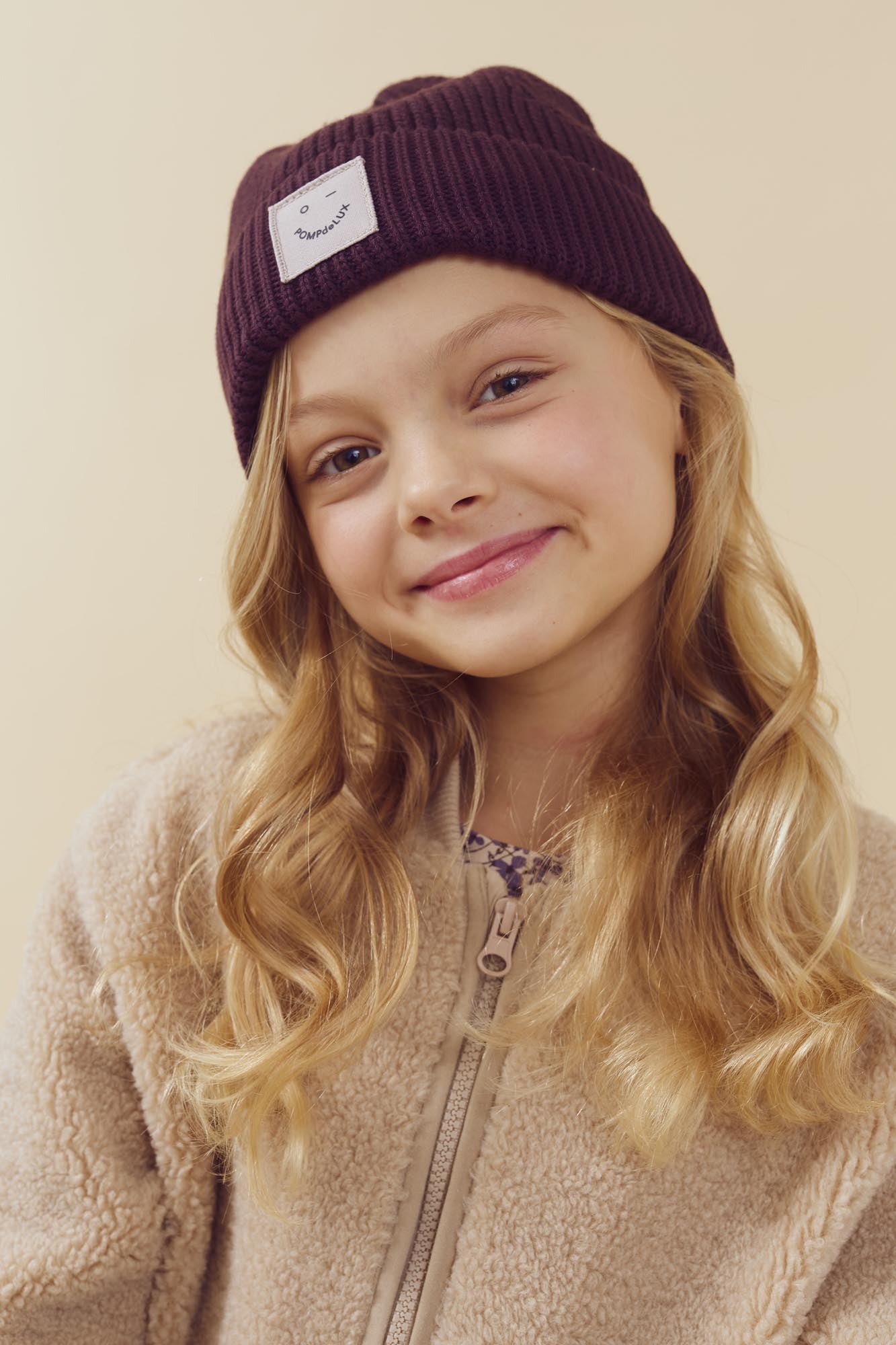 A child with long blonde hair smiles, wearing a maroon beanie with a "POMPdeLUX" label, and a fluffy beige jacket, set against a plain light background.