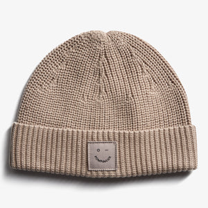 A beige knit beanie with a folded brim displays a small rectangular patch reading "pompdelux" and a minimalist face logo. It's placed on a plain white background.