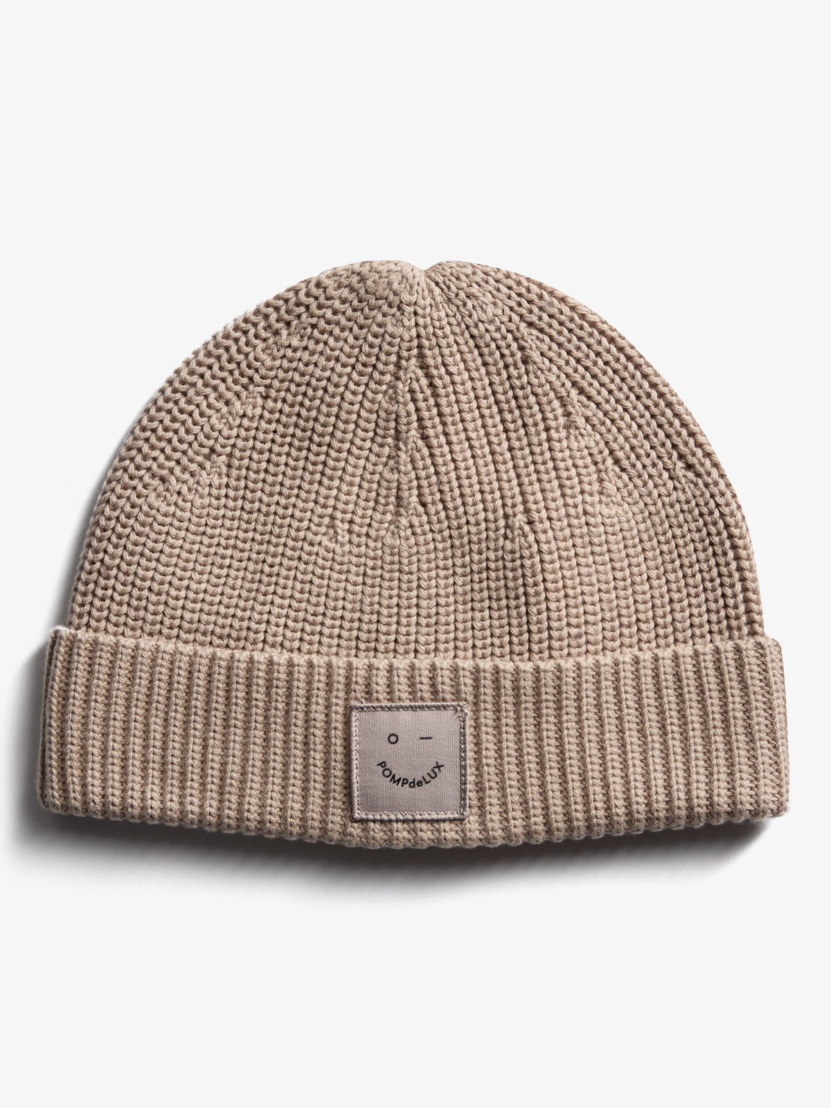 A beige knit beanie with a folded brim displays a small rectangular patch reading "pompdelux" and a minimalist face logo. It's placed on a plain white background.