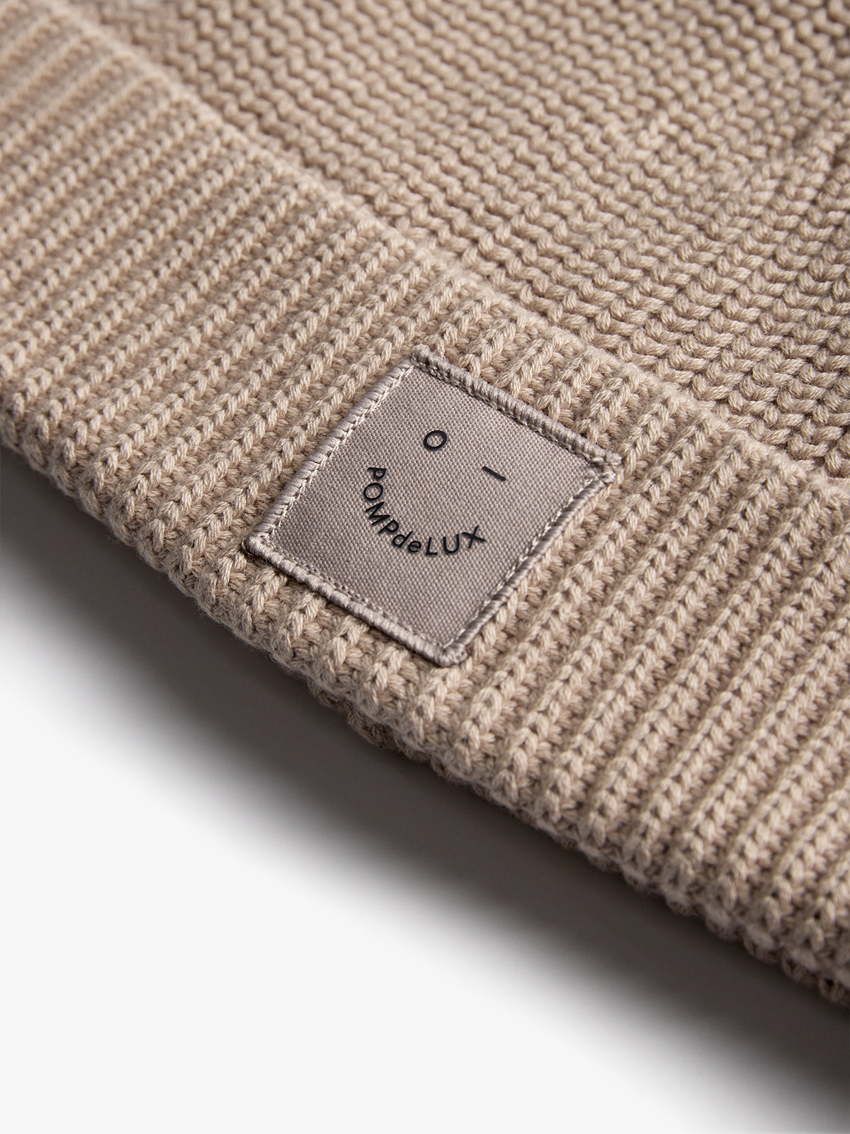 A beige knit hat with a woven texture features a small patch displaying a simple face and the text "POMP de LUX." The hat fills the foreground.