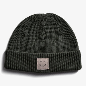A knitted dark green beanie displays a small patch with a smiley face and the text "POMPdeLUX" against a plain white background.