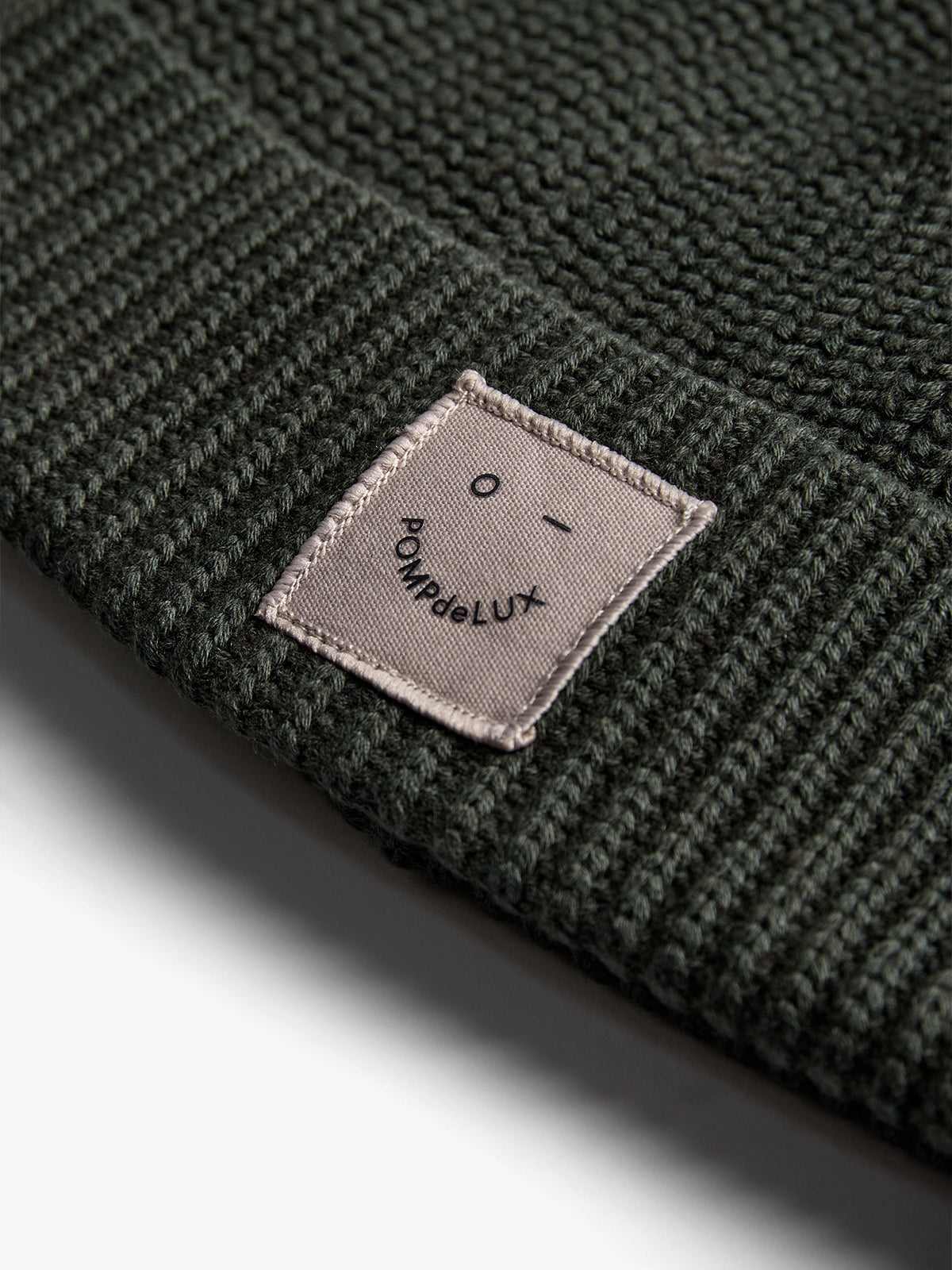 A dark green knitted beanie displays a sewn beige label with a simple face design and the text "POMPdeLUX." The beanie rests against a neutral background.