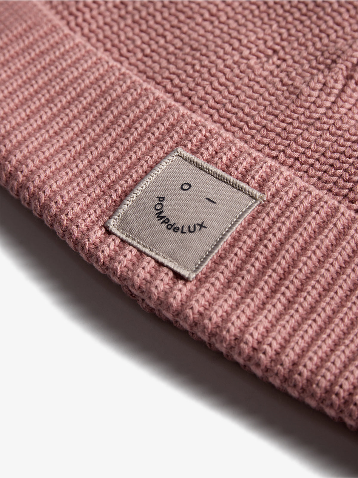 A pink knit beanie displays a small fabric label featuring a simple smiley face and the text "POMPdeLUX" in a minimalistic, gray design.